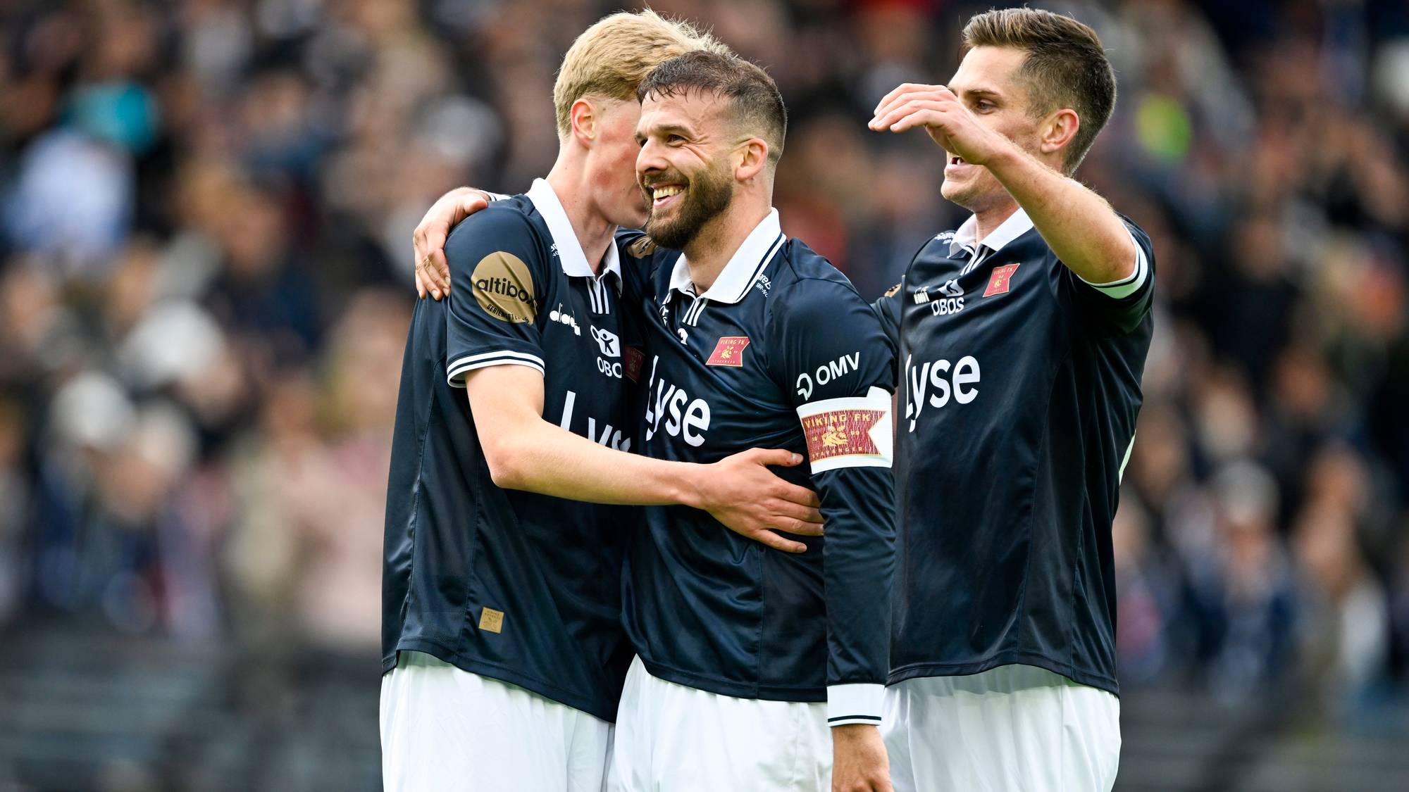 Tre fotballspillere i mørke drakter omfavner hverandre. De smiler og ser ut til å feire. I bakgrunnen skimtes et publikum på en tribun. (Bildebeskrivelsen er laget av en KI-tjeneste)