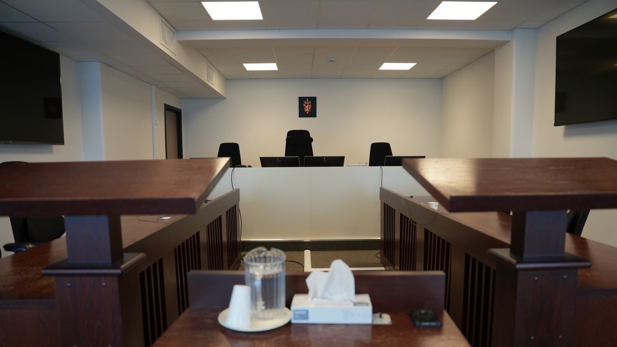 Et rettssalsinteriør med tre tomme dommerstoler i bakgrunnen, bak et hvitt panel. Foran dommerstablene ses det et norsk riksvåpen. I forgrunnen er det et bord med en vannkaraffel og en papirserviettboks. (Bildebeskrivelsen er laget av en KI-tjeneste) FOTO: Oskar Rennedal/NRK