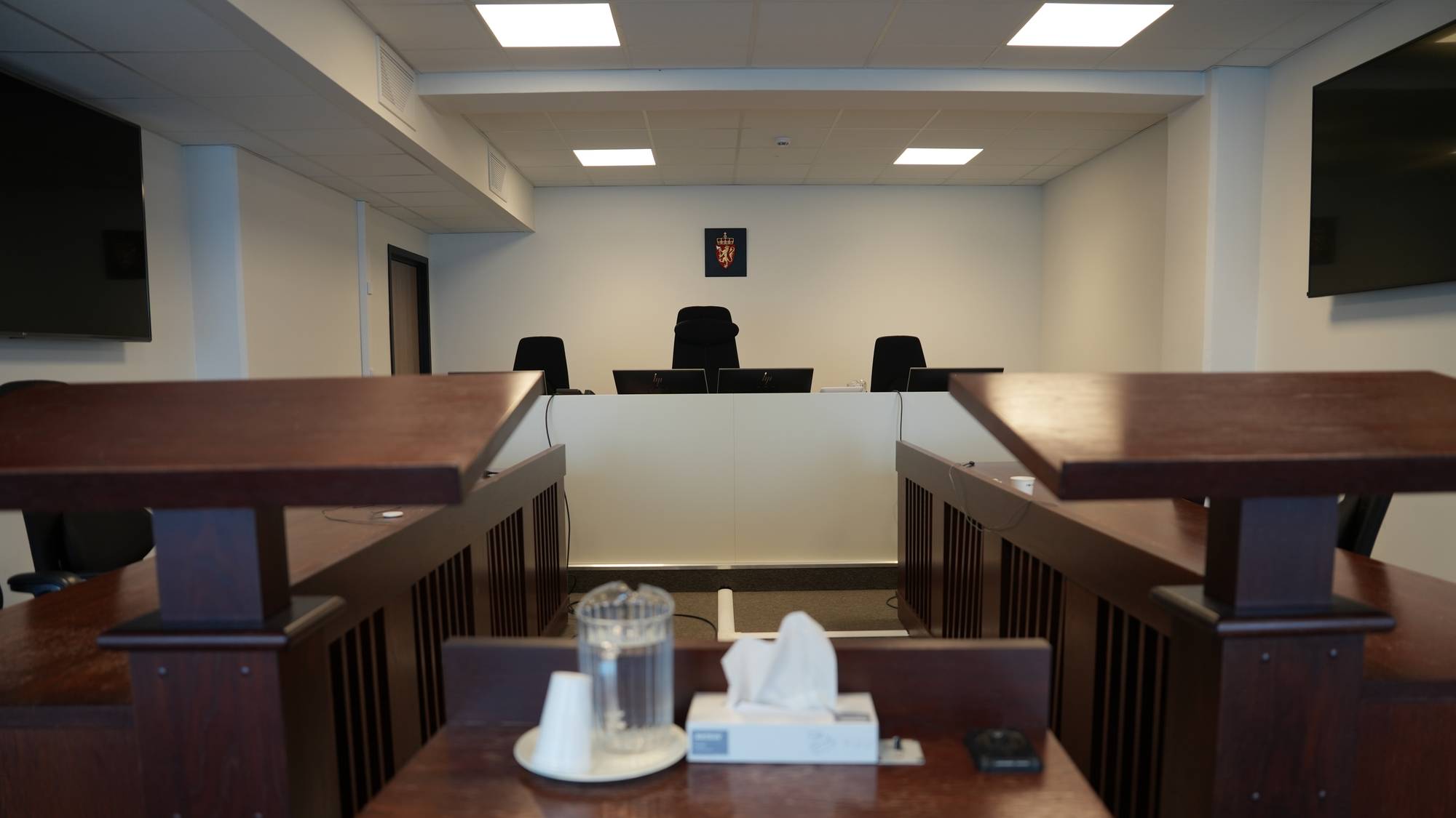 Et rettssalsinteriør med tre tomme dommerstoler i bakgrunnen, bak et hvitt panel. Foran dommerstablene ses det et norsk riksvåpen. I forgrunnen er det et bord med en vannkaraffel og en papirserviettboks. (Bildebeskrivelsen er laget av en KI-tjeneste)