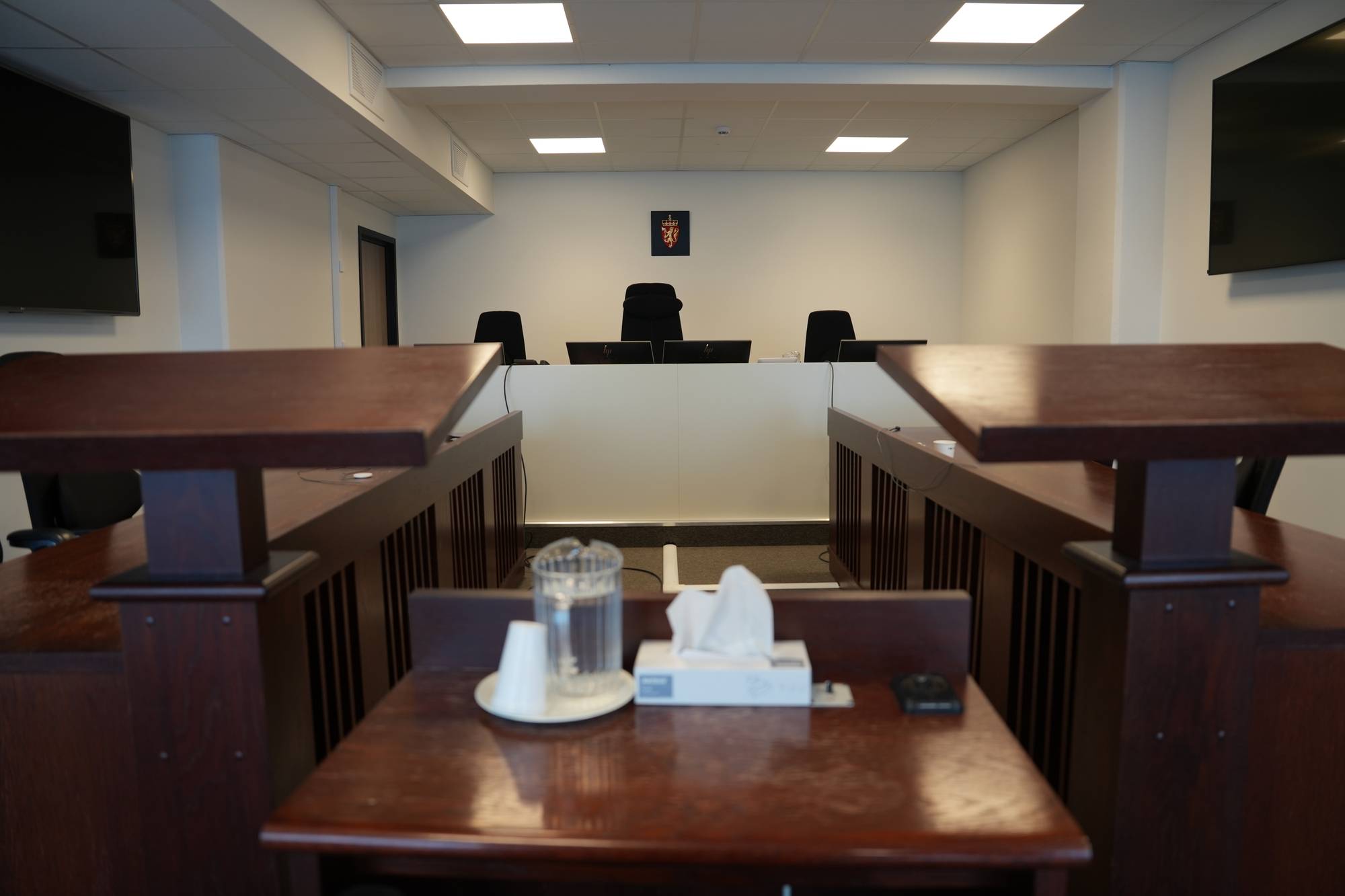 Et rettssalsinteriør med tre tomme dommerstoler i bakgrunnen, bak et hvitt panel. Foran dommerstablene ses det et norsk riksvåpen. I forgrunnen er det et bord med en vannkaraffel og en papirserviettboks. (Bildebeskrivelsen er laget av en KI-tjeneste)