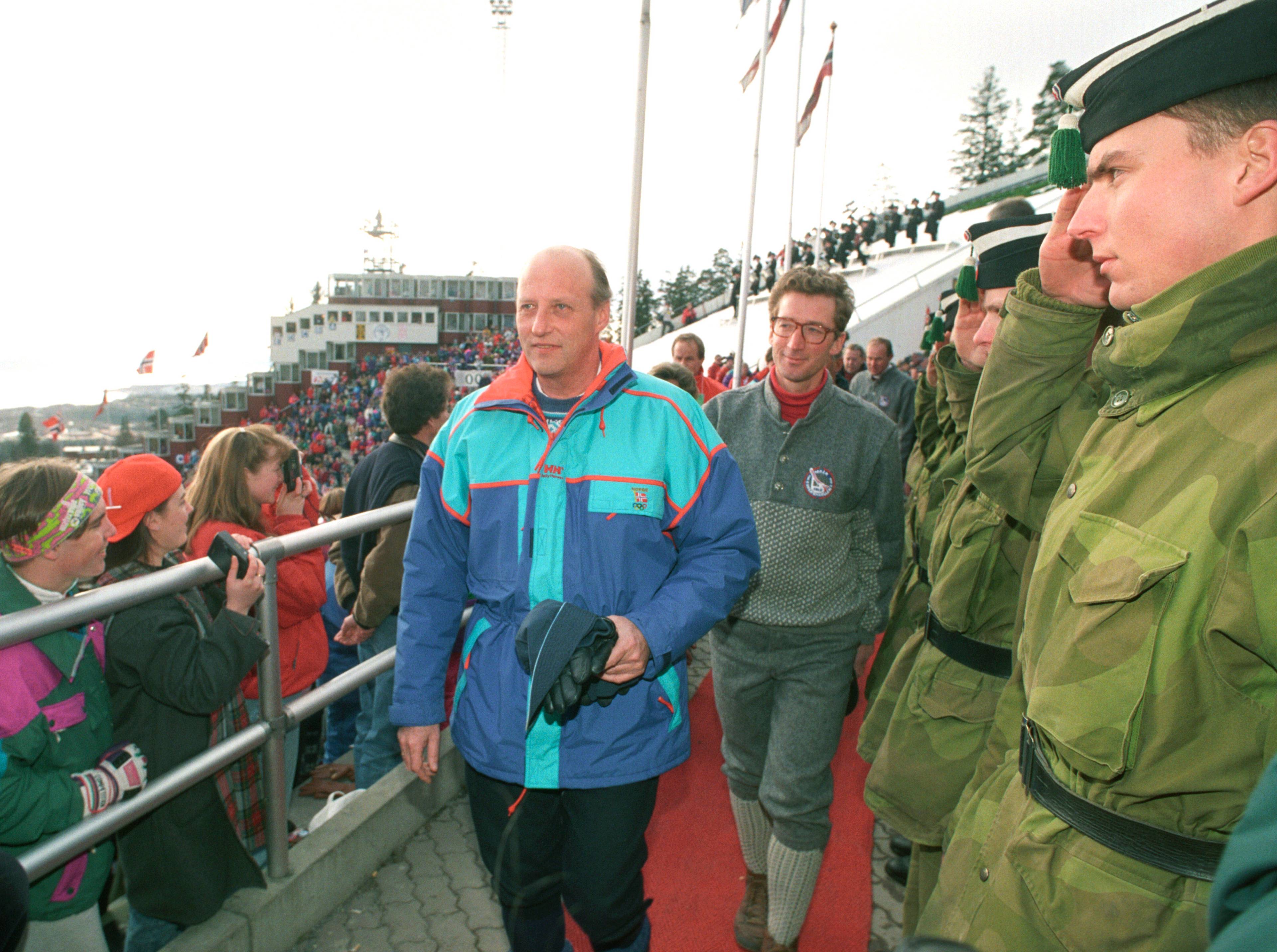 Kong Harald går på rød løper forbi æresvakt i militæruniform ved hoppbakke. Tilskuere ser på. (Bildebeskrivelsen er laget av en KI-tjeneste)