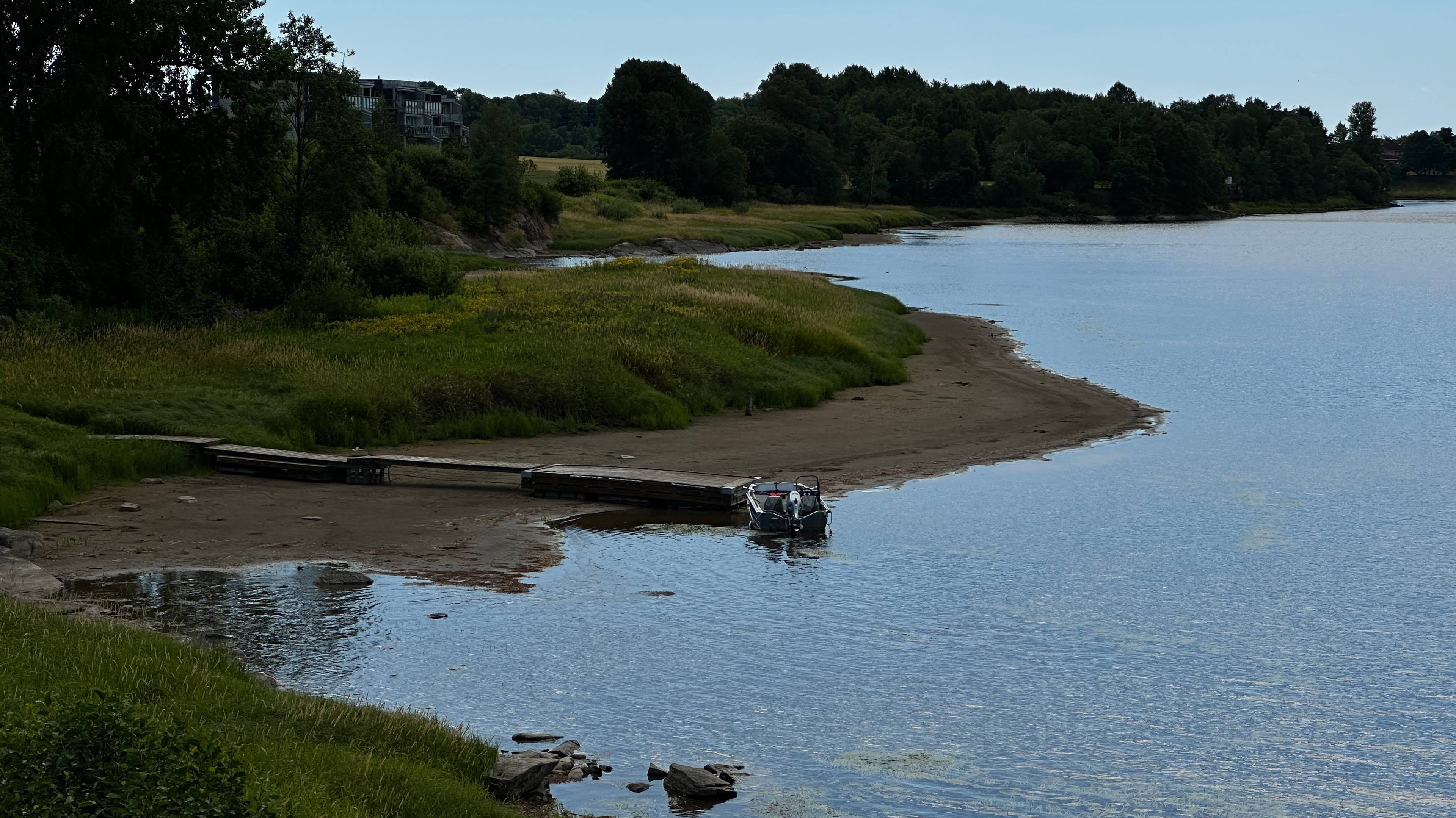 En liten båt ligger fortøyd til en trebrygge ved en strandlinje omgitt av skog og grøntareal. (Bildebeskrivelsen er laget av en KI-tjeneste)