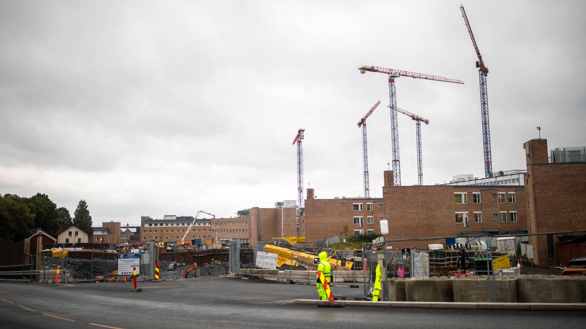 Byggeplass med flere tårnkraner og teglsteinsbygg i bakgrunnen under en overskyet himmel. (Bildebeskrivelsen er laget av en KI-tjeneste)