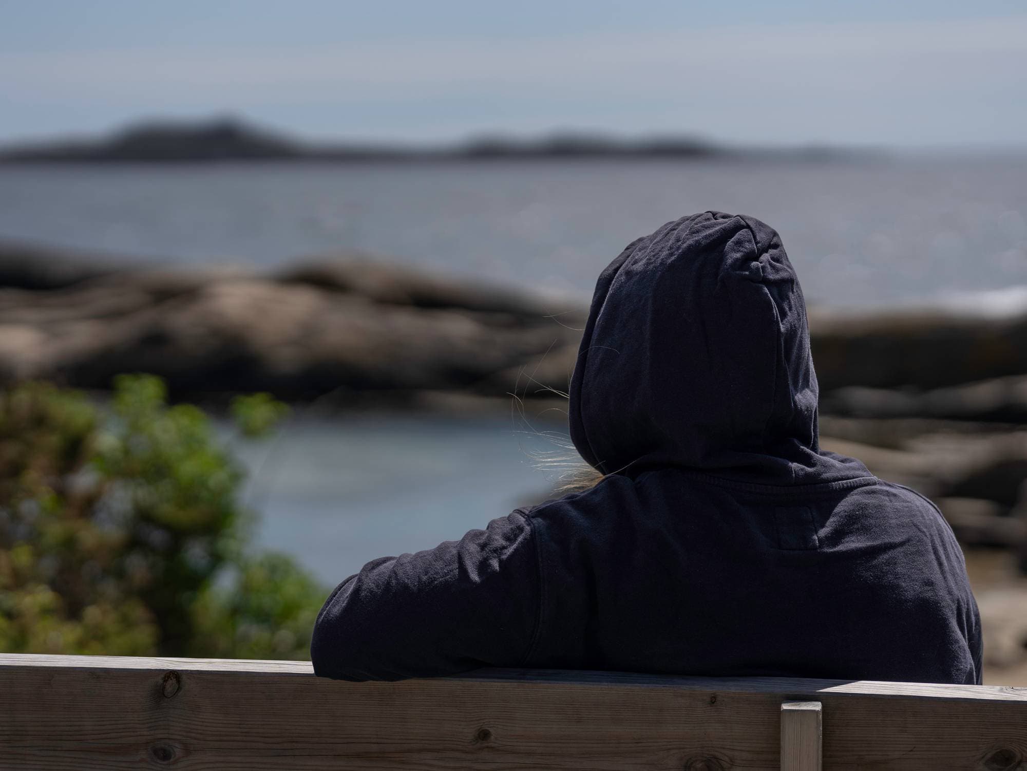 kvinne sitter på en benk med ryggen til. Hun har på svart hettegenser. Hun ser utover mot havet. 