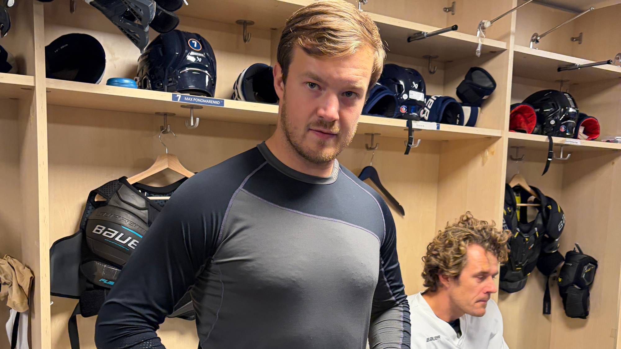 Ishockeyspiller i garderobe med utstyr i bakgrunnen. En annen spiller sitter på en benk. (Bildebeskrivelsen er laget av en KI-tjeneste)