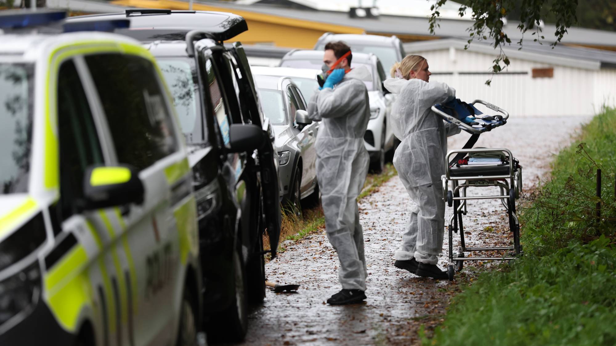 Krimteknikere i hvite drakter ved politibiler på et åsted. En person håndterer en båre. (Bildebeskrivelsen er laget av en KI-tjeneste)