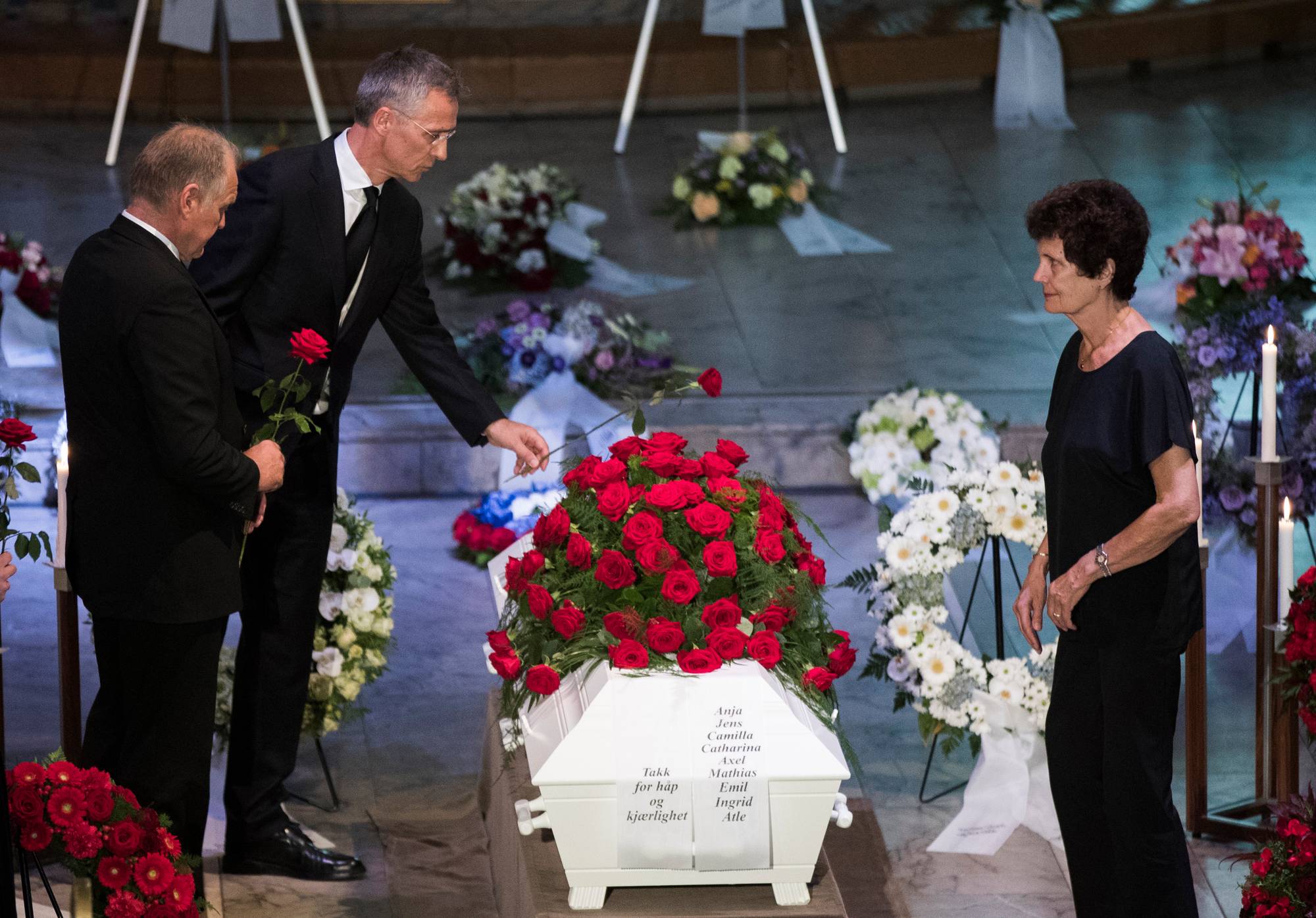 Jens Stoltenberg legger en rød rose på en hvit kiste, som er dekket av mange røde roser. Ved siden av står en annen mann i dress, og Anja Breien i svart kjole ser mot kisten. Det er blomster og lys rundt kisten.