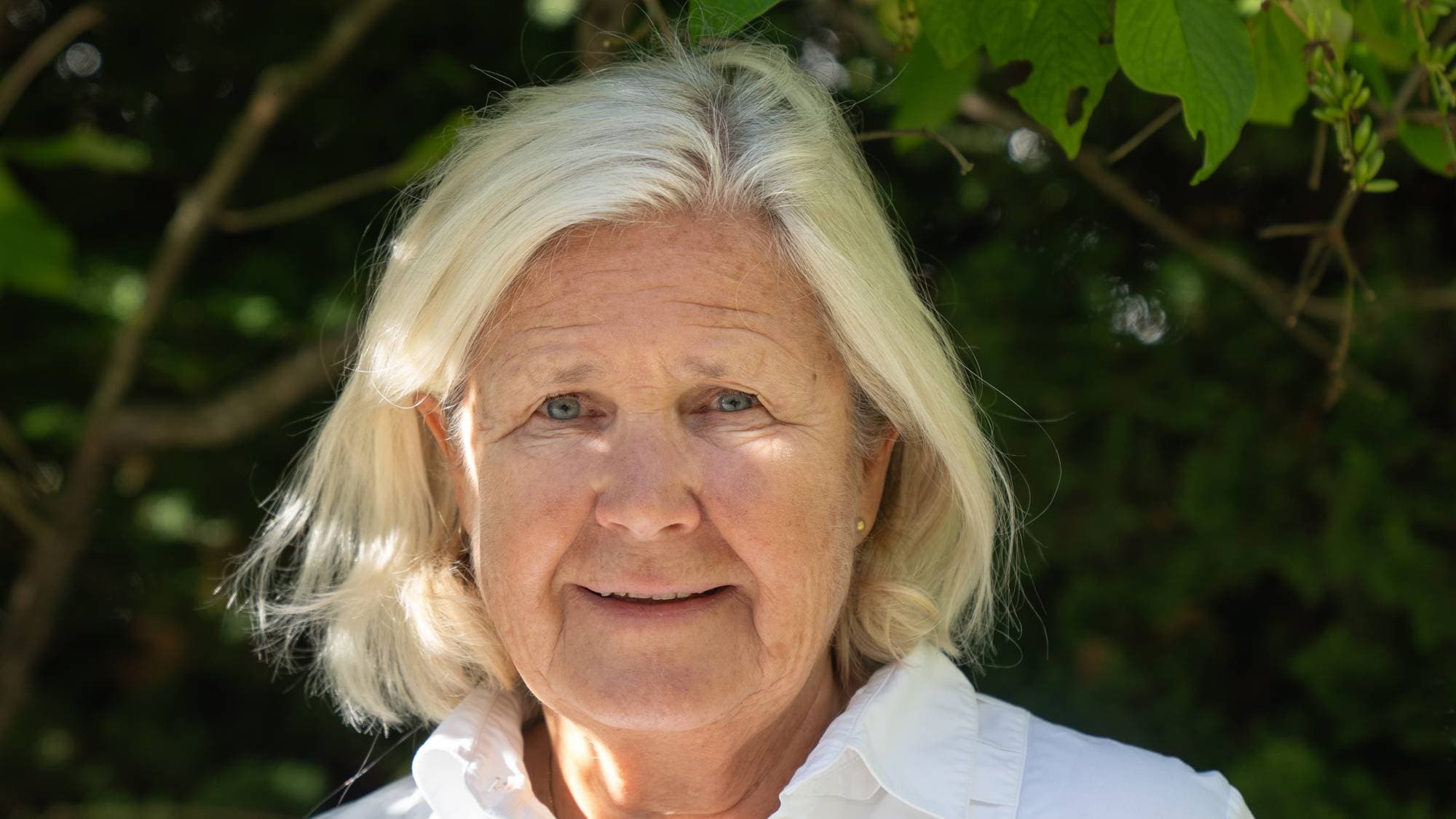 Anette Hylen Ranhoff stiller til portrettbildet i hvit skjorte med en grønn plantevegg i bakgrunn