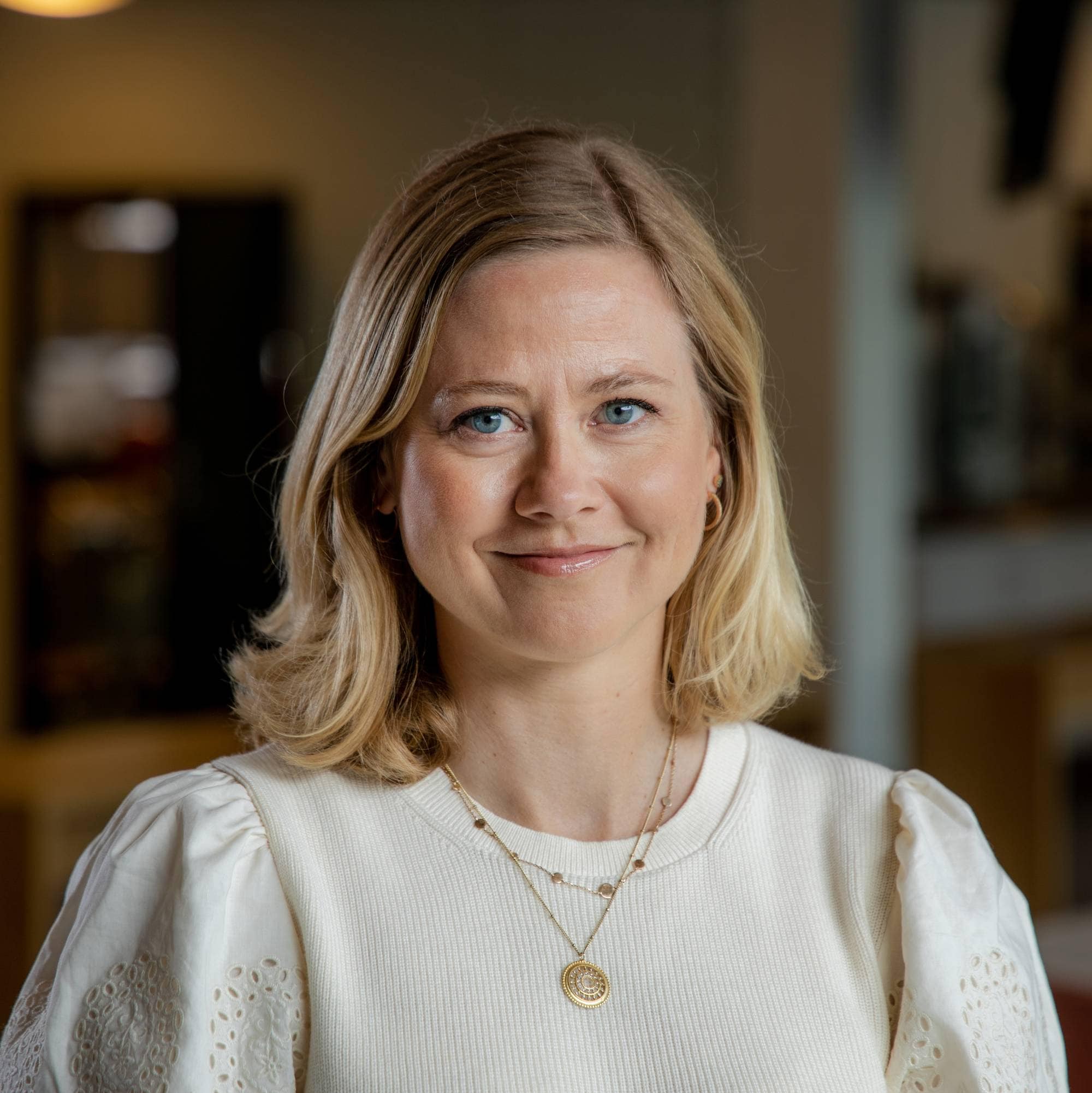 Portrett av en lyshåret kvinne i en lys overdel, avbildet foran en uskarp kontorbakgrunn. (Bildebeskrivelsen er laget av en KI-tjeneste)