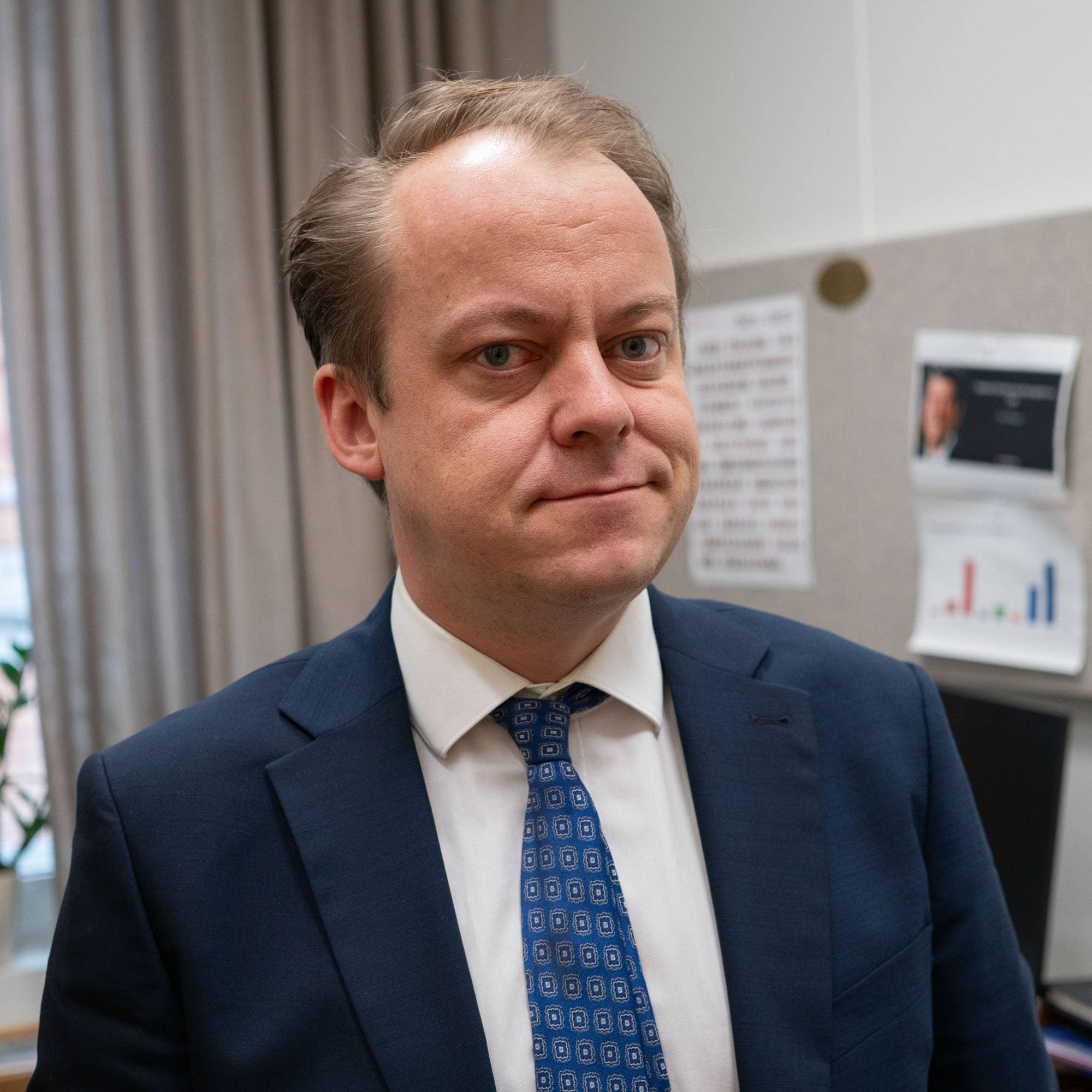 En mann i dress og slips ser mot kamera. Han står inne på et kontor.