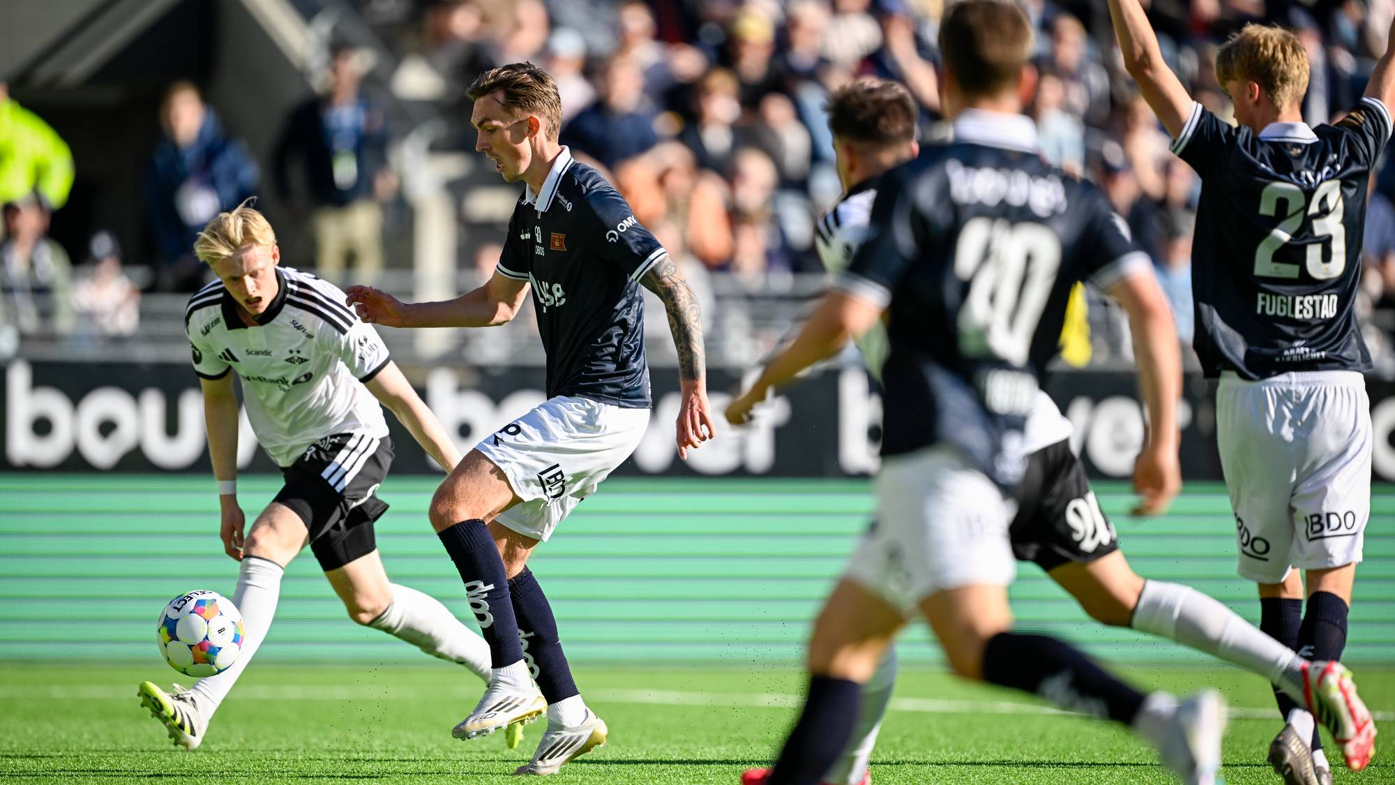 Fotballspillere i mørke draktkonkurrerer om ballen på en kunstgressbane. En spiller i hvit drakt sparker til ballen. Tilskuere sitter på tribunen i bakgrunnen. (Bildebeskrivelsen er laget av en KI-tjeneste)