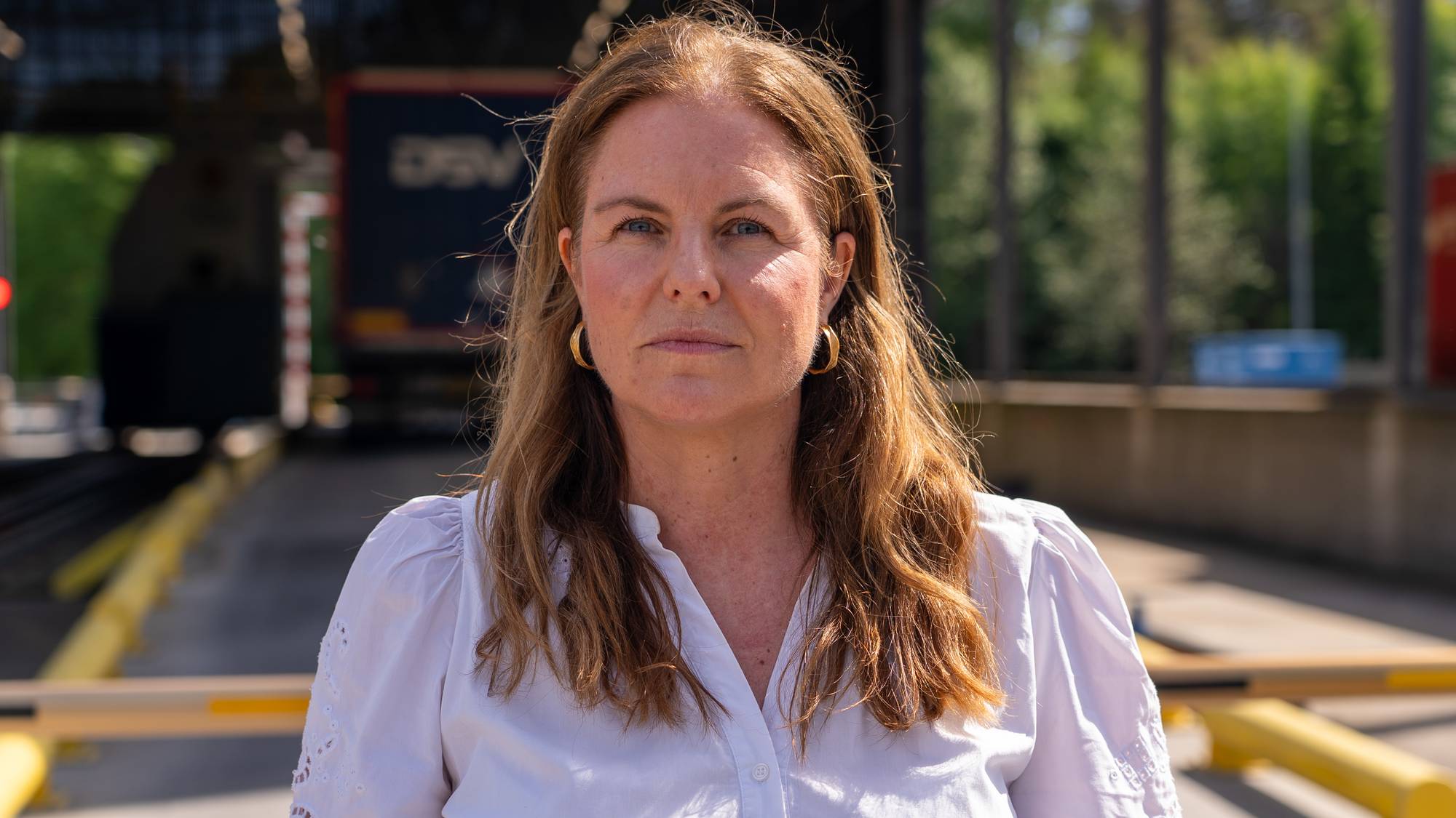 En kvinne i hvit skjorte med store gullsirkeløredobber ser mot kameraet. Bak henne ses en lastebil med teksten "DSV" og en bygning med vinduer. Omgivelsene er utendørs, med sollys. (Bildebeskrivelsen er laget av en KI-tjeneste)
