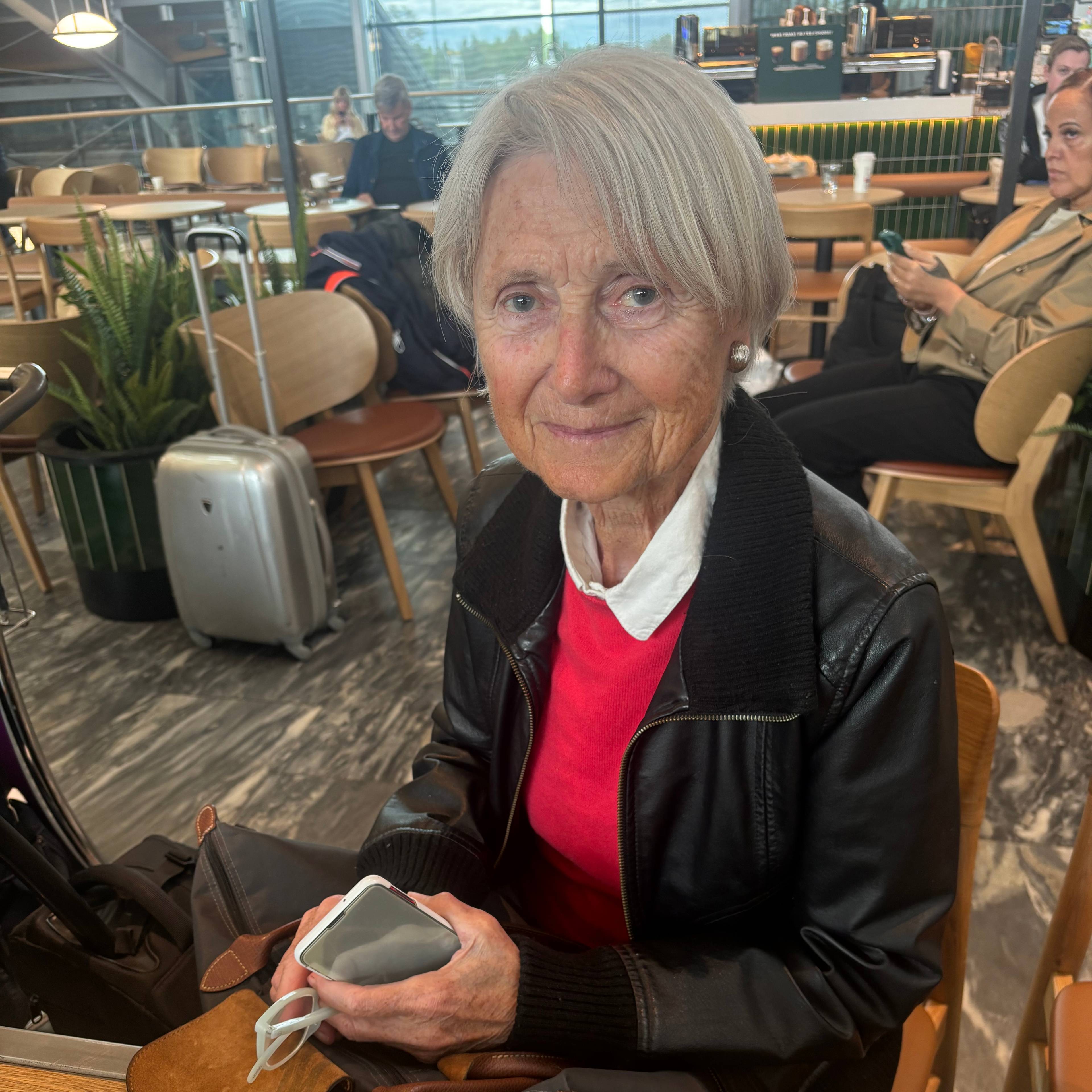 En eldre kvinne sitter på en flyplass. Hun har på seg en rød genser og en svart skinnjakke.