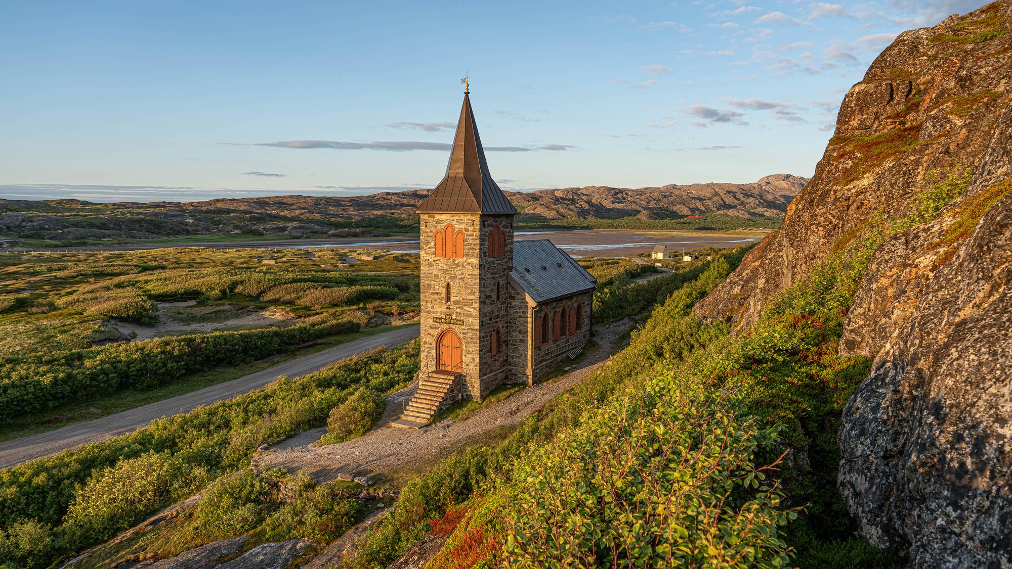 Et steinkirke med spir står på en skråning. En grusvei går forbi kirken. Kirken er omgitt av grønt landskap med fjell i bakgrunnen. (Bildebeskrivelsen er laget av en KI-tjeneste)