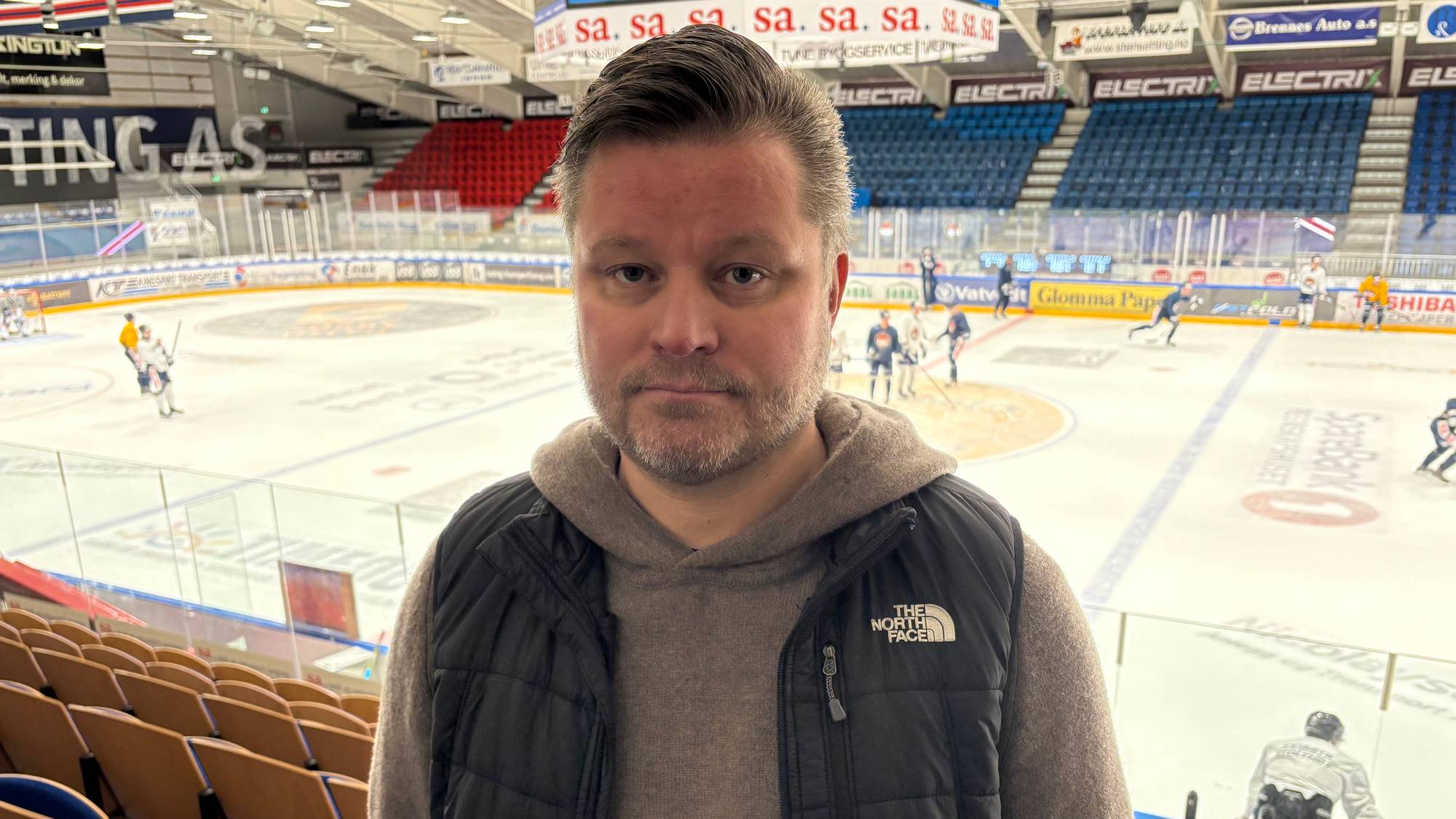 Mann i 40-årene står på tribunen i en ishall med spillere på isen i bakgrunnen. (Bildebeskrivelsen er laget av en KI-tjeneste)