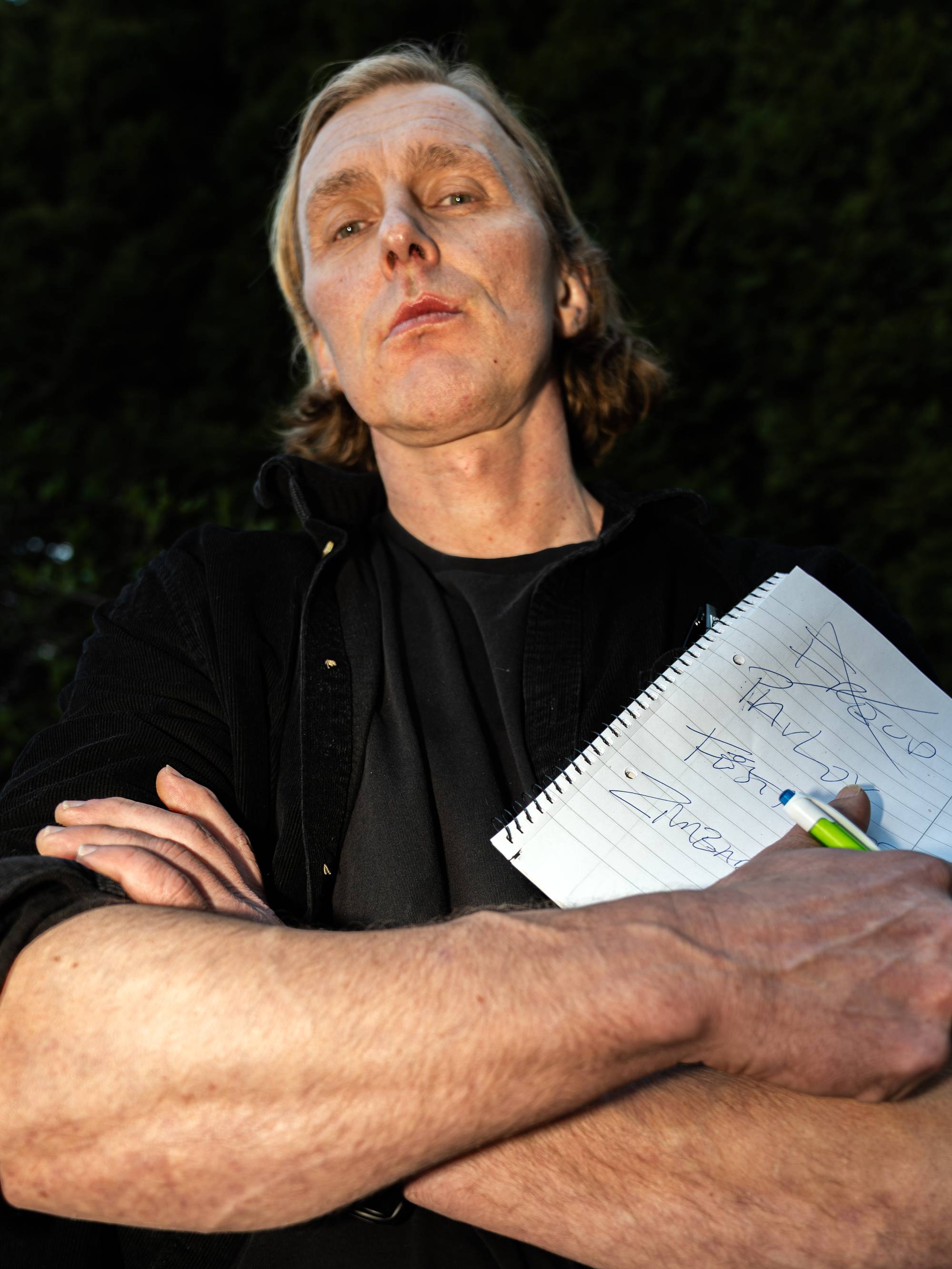 En mann med blondt hår og skjeggstubber holder en notatblokk med pen. Han har på seg en svart skjorte og jakke. Mannen står foran en mørk bakgrunn. (Bildebeskrivelsen er laget av en KI-tjeneste)