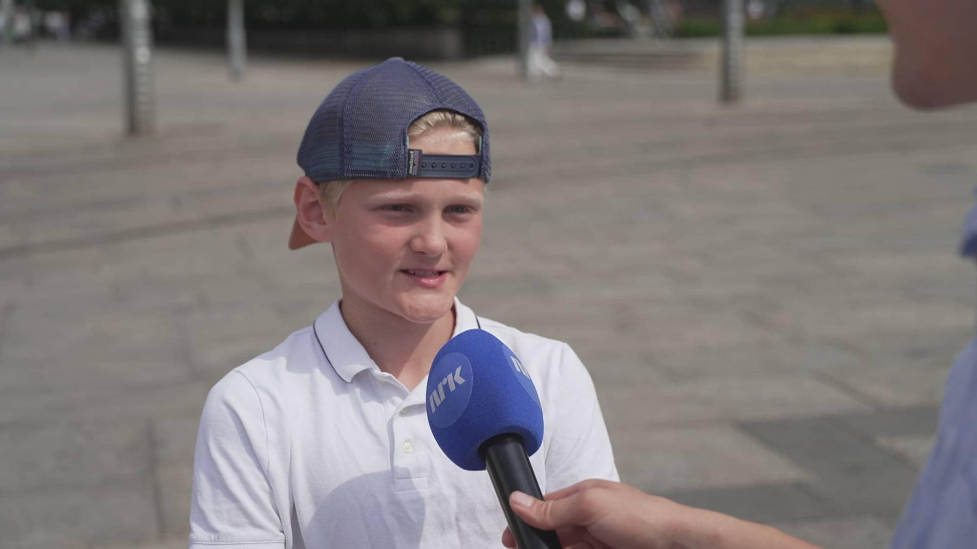 En ung gutt med caps intervjues av en person som holder en NRK-mikrofon. Utendørs bakgrunn. (Bildebeskrivelsen er laget av en KI-tjeneste)