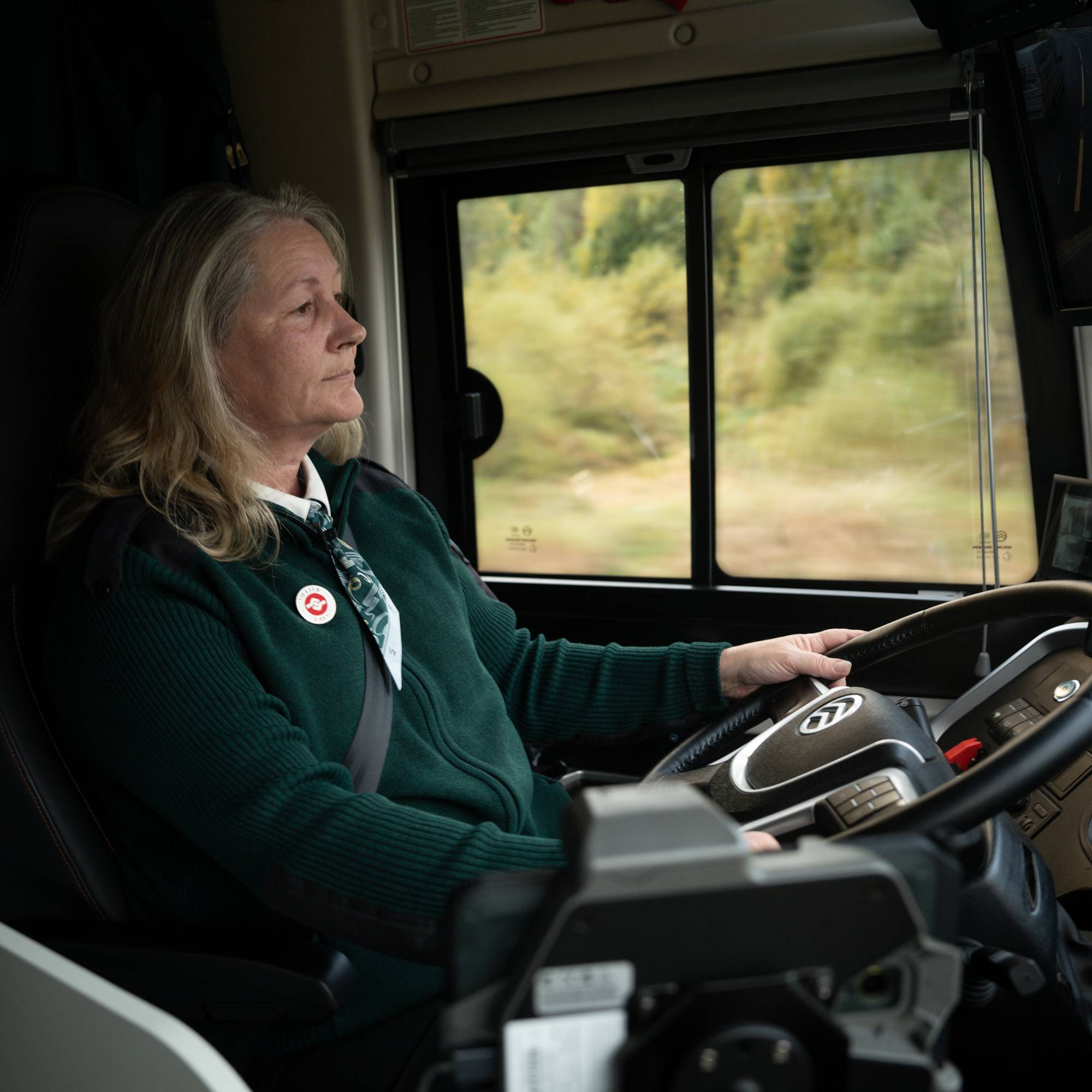 En kvinne i grønn uniform sitter bak rattet og kjører en buss.