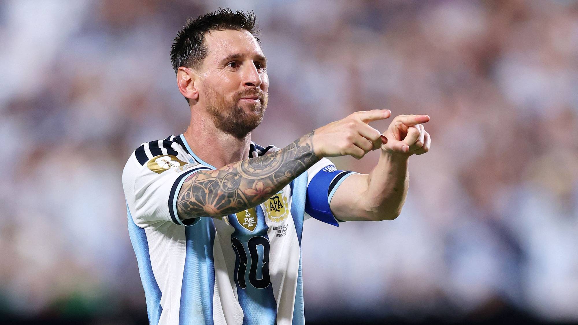 Lionel Messi peker med begge hender under en fotballkamp. Han har på seg Argentinas drakt med nummer 10. Bak ham er en uskarpt bakgrunn med publikum. (Bildebeskrivelsen er laget av en KI-tjeneste)