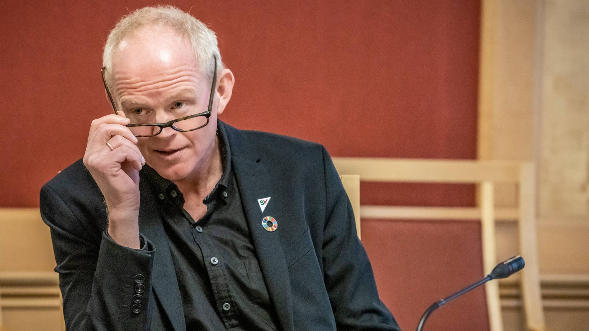 En person sitter ved et bord i en møtesal, iført en svart dressjakke og skjorte. Personen ser ut til å være i tankefull positur, og med den ene hånden tar han av brillene sine. På bordet ligger flere dokumenter og en notisbok. Møtesalen har en rød vegg og en mikrofon foran personen. (Bildebeskrivelsen er laget av en KI-tjeneste)