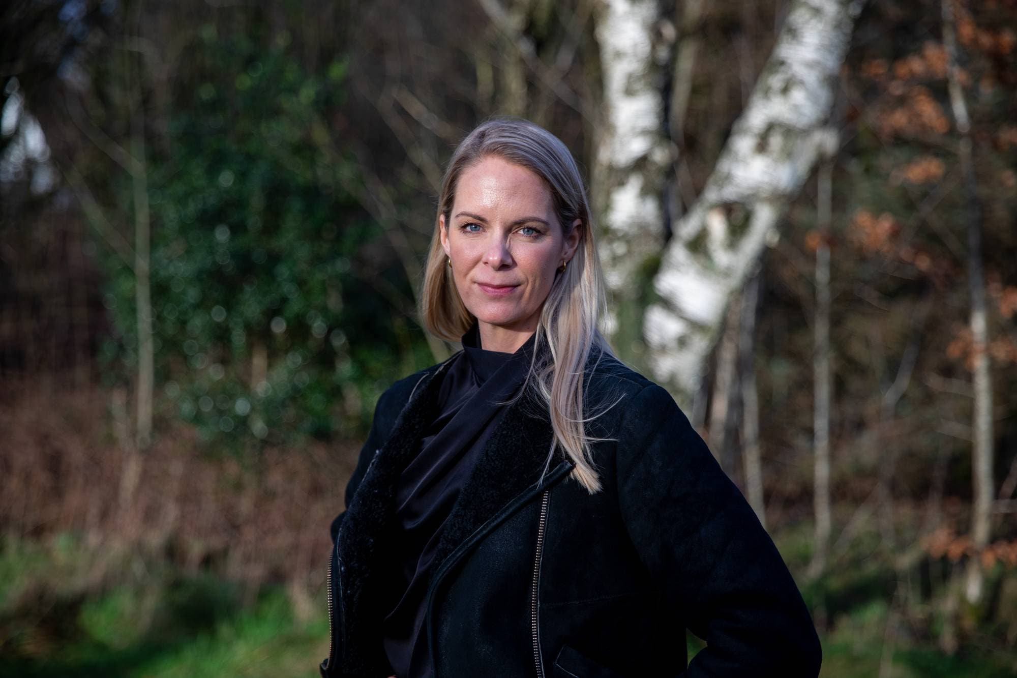 Kvinne med blondt hår og i svart jakke står utendørs med trær i bakgrunnen