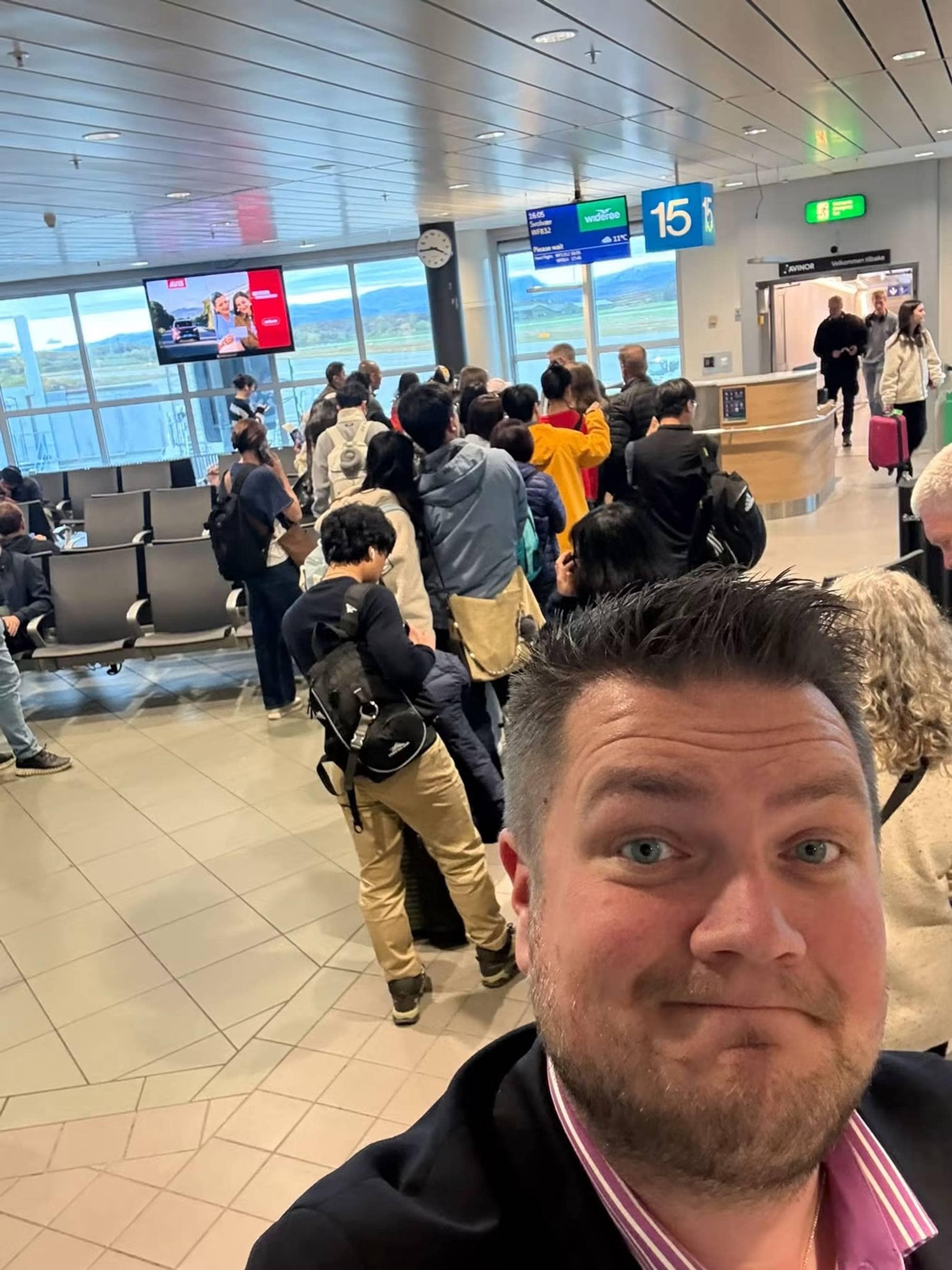 En mann tar en selfie i en flyplassgate mens reisende står i kø ved en utgang. (Bildebeskrivelsen er laget av en KI-tjeneste)