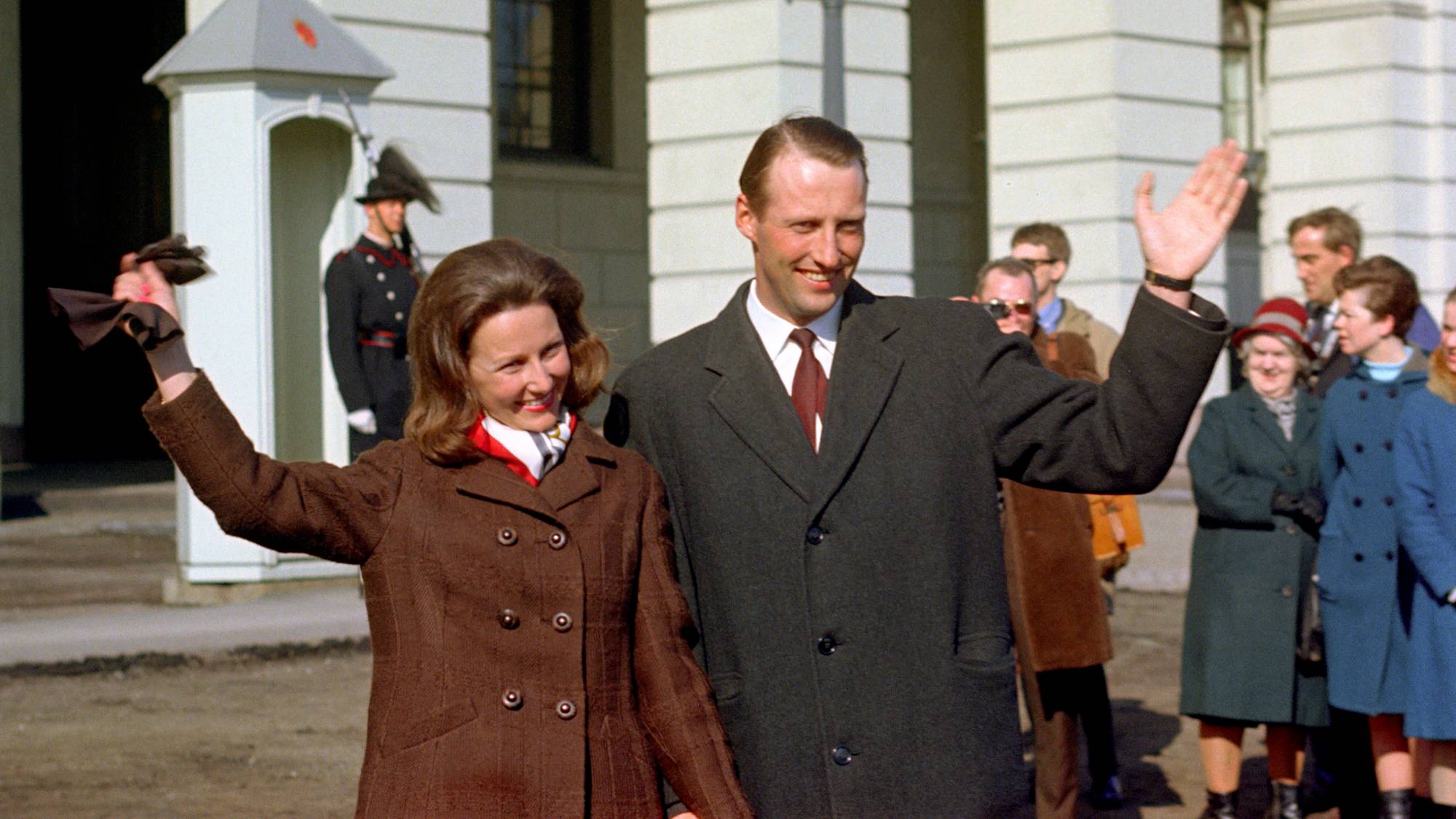 Kong Harald og dronning Sonja vinker foran Slottet med en gardist i bakgrunnen. (Bildebeskrivelsen er laget av en KI-tjeneste)