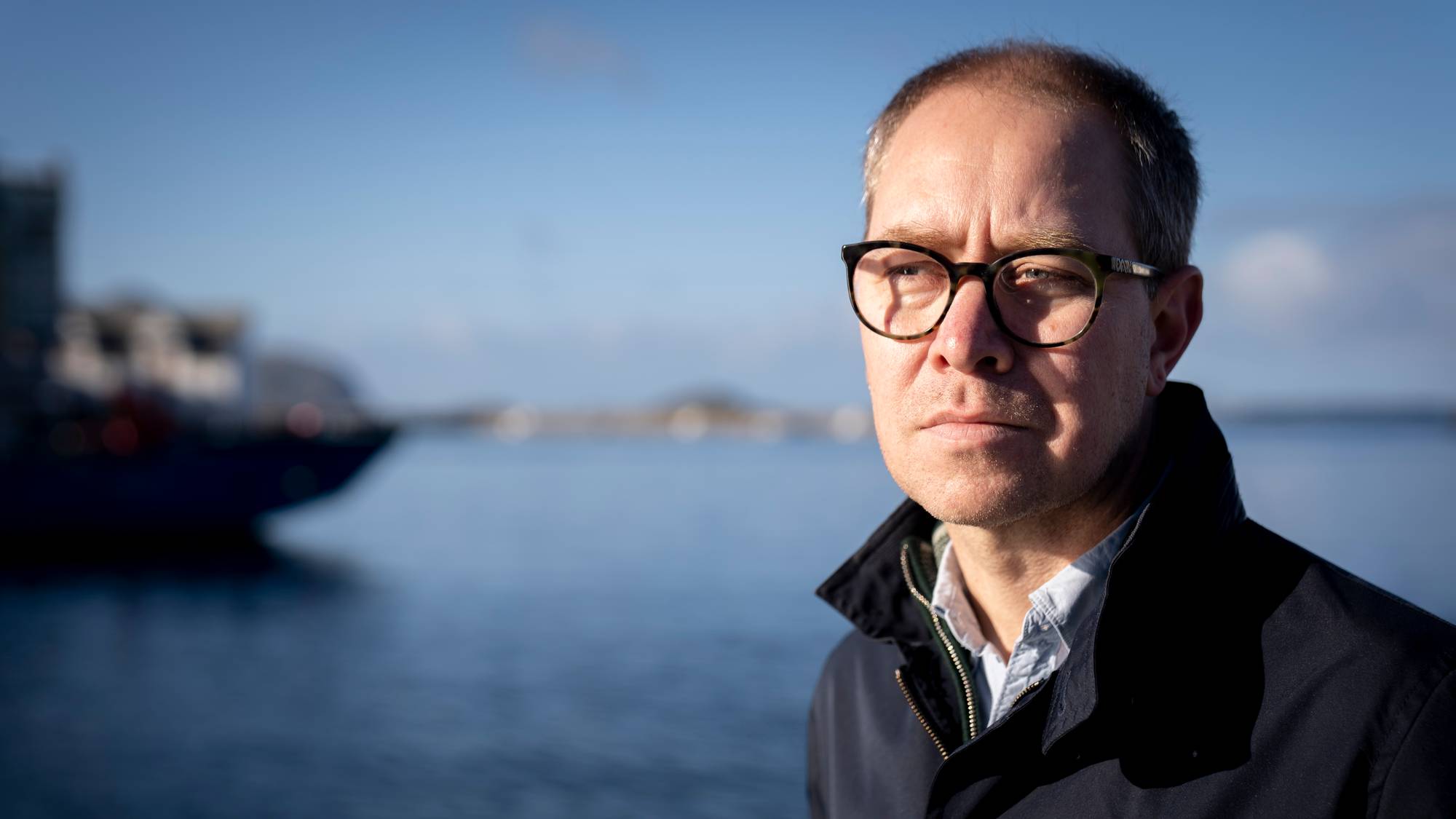 Sverre Johansen står med ein blå frakk og ser alvorleg på noko på sida av kameraet. Bak han ser vi havet, ein molo og ein båt til kai. Himmelen er blå med nokre skyer. Johansen har på seg briller og sola kastar harde skuggar.
