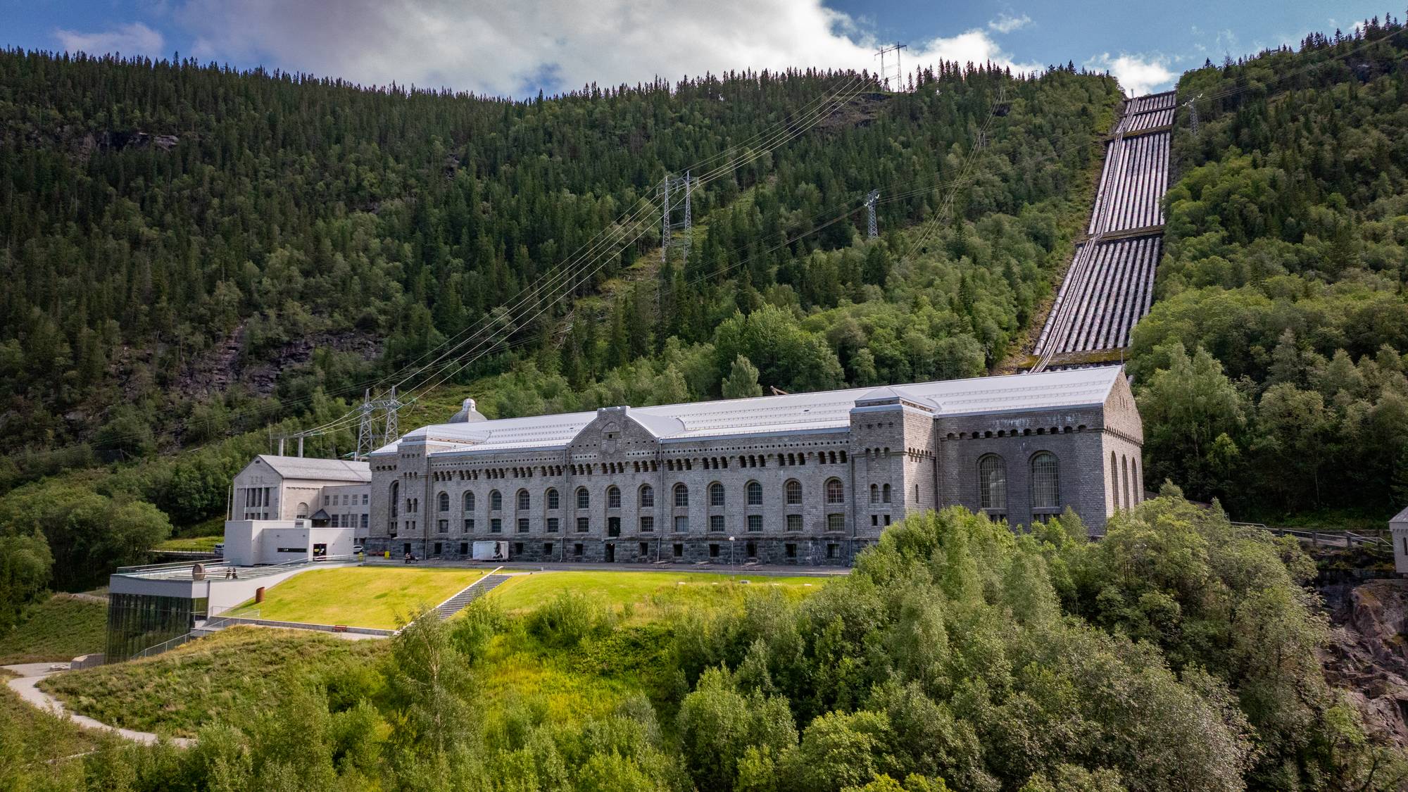Vemork kraftstasjon i et kupert landskap med rørgater oppover fjellsiden. (Bildebeskrivelsen er laget av en KI-tjeneste)