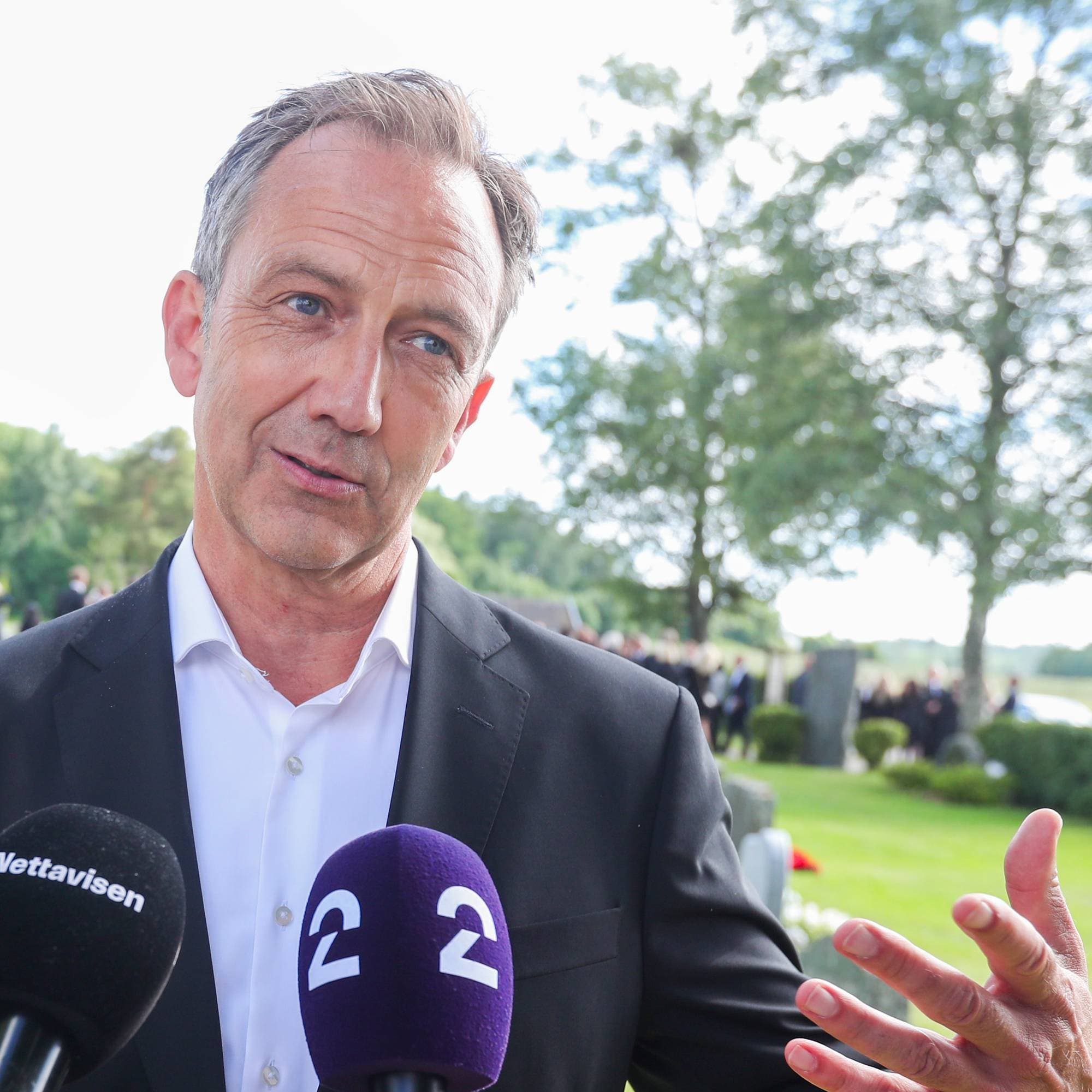 Kvinne og mann i svarte klær blir intervjuet ved en kirkegård. (Bildebeskrivelsen er laget av en KI-tjeneste)