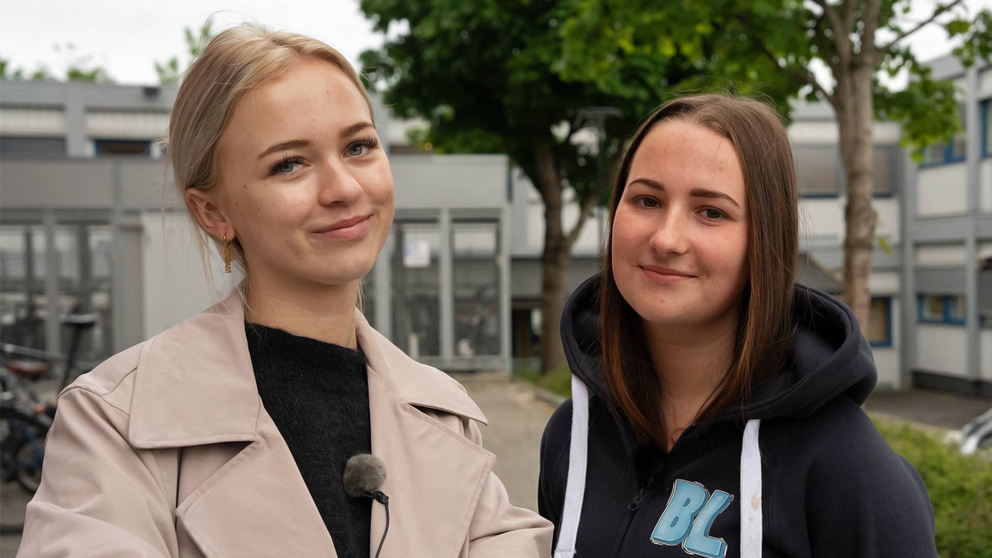 En blond jente med hestehale og beige jakke står ved siden av en annen ung jente med brunt hår og russegenser.