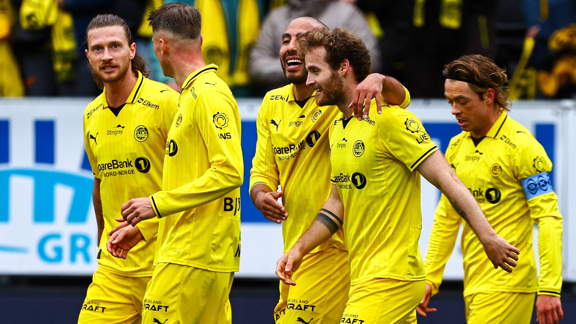 Fem fotballspillere i gule drakter feirer på en gressbane. Spilleren lengst til høyre har et armbind. I bakgrunnen er det tilskuere og reklameskilt. (Bildebeskrivelsen er laget av en KI-tjeneste)