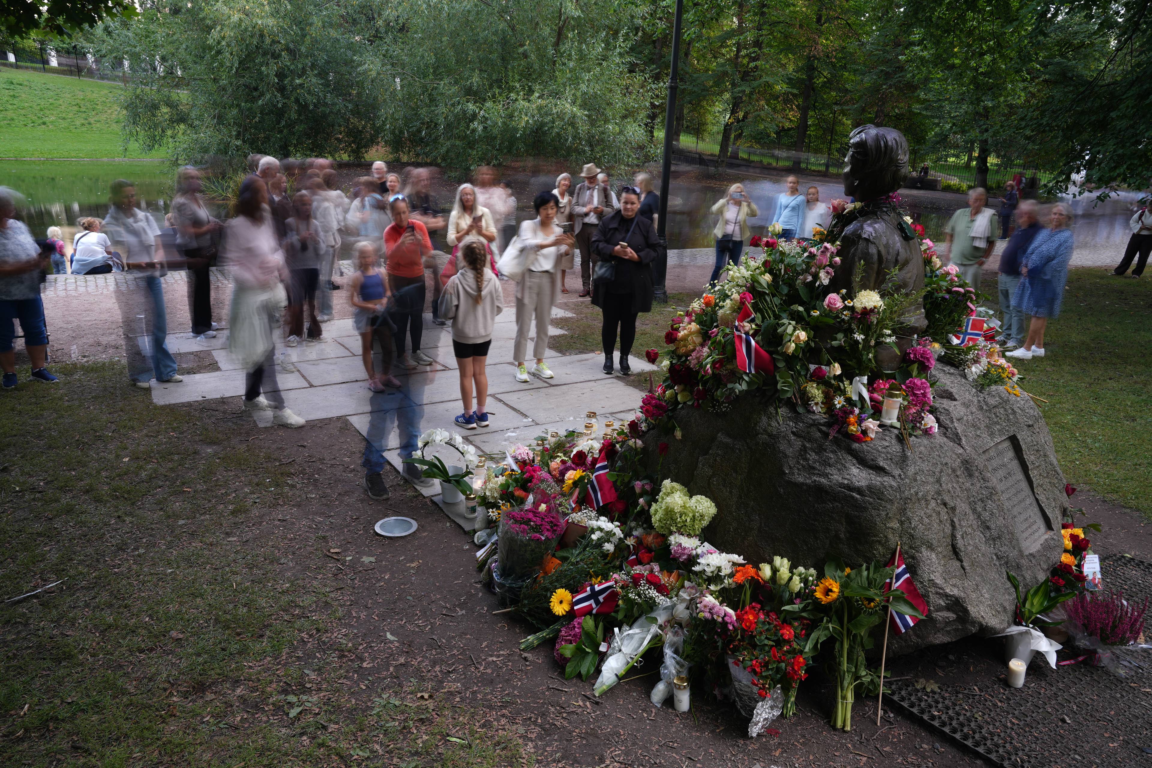 Folk legger ned blomster ved en byste i en park, omgitt av flagg og tente lys. (Bildebeskrivelsen er laget av en KI-tjeneste)