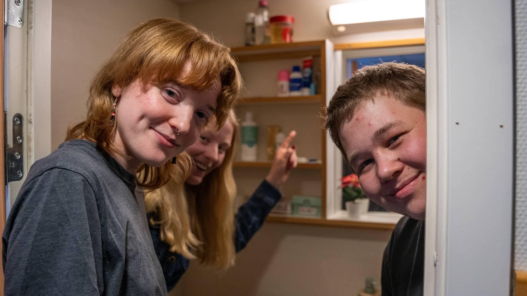 Tre unge mennesker kikker ut av en baderomsdør og smiler. 