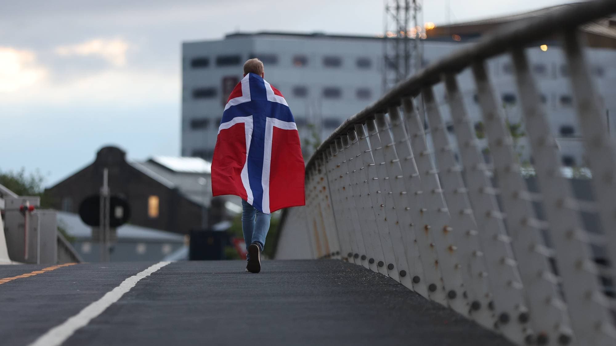 Person går over en bro med et norsk flagg svøpt rundt skuldrene. Bygninger i bakgrunnen. (Bildebeskrivelsen er laget av en KI-tjeneste)