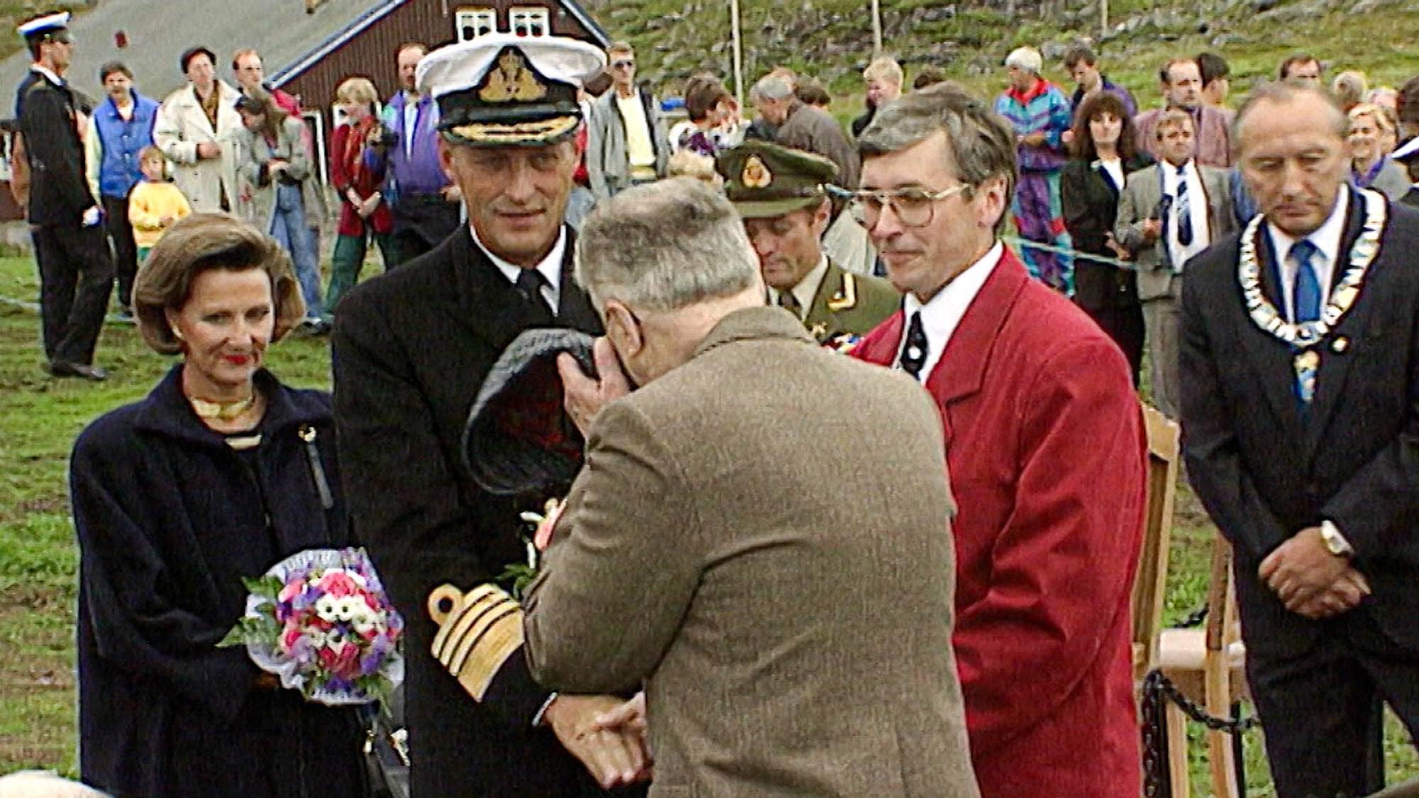 Kong Harald og Dronning Sonja ved en seremoni utendørs med uniformerte personer og en ordfører. (Bildebeskrivelsen er laget av en KI-tjeneste)