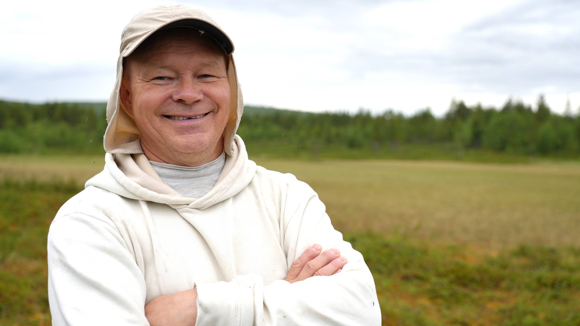 En mann med hvit hettegenser og hvit kaps ser inn i kamera med hendene i korst og smiler. Han står i en myr.