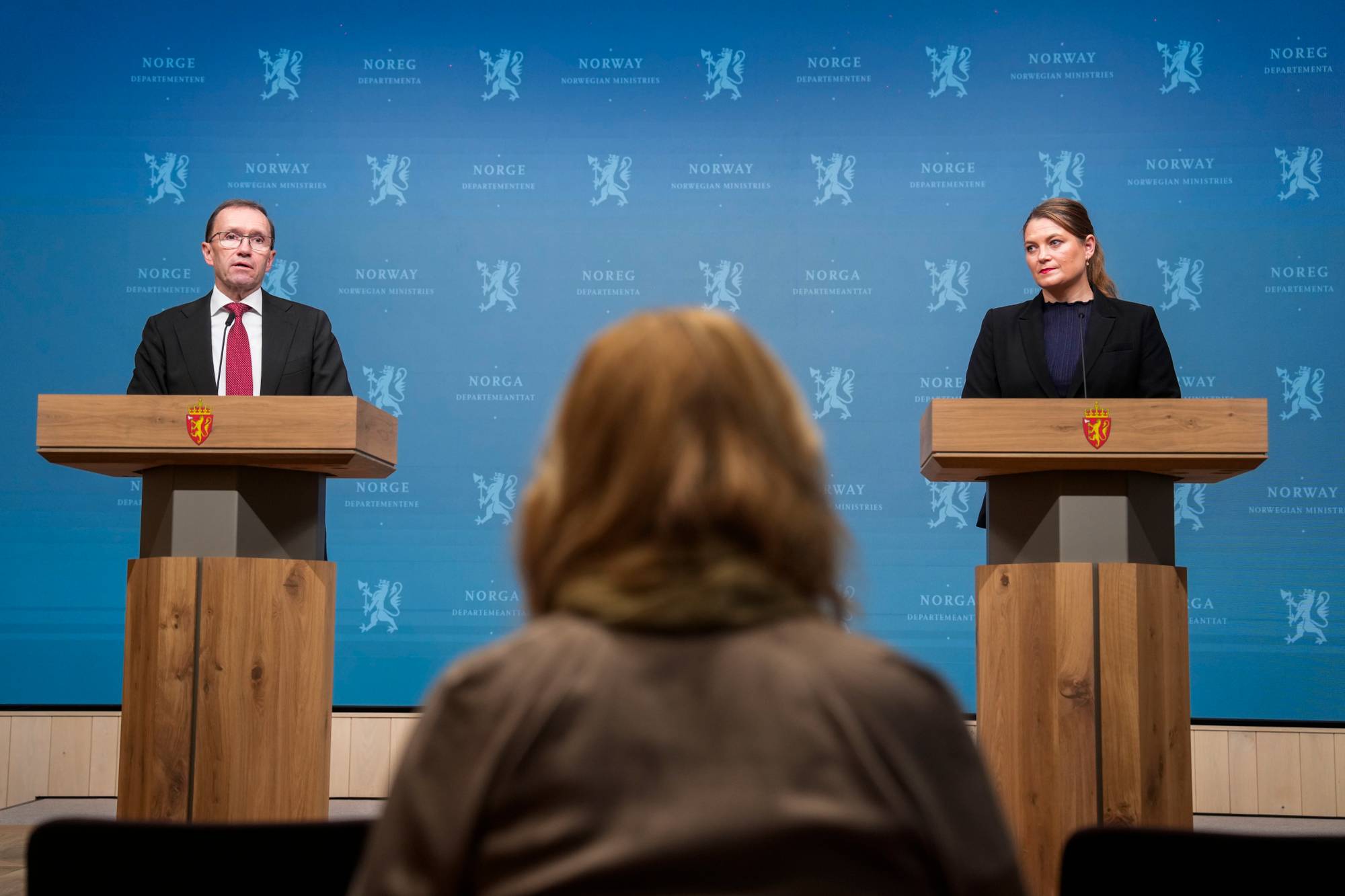 En mann og en kvinne står bak hver sin talerstol under en pressekonferanse. En blå vegg med regjeringslogo er bak dem