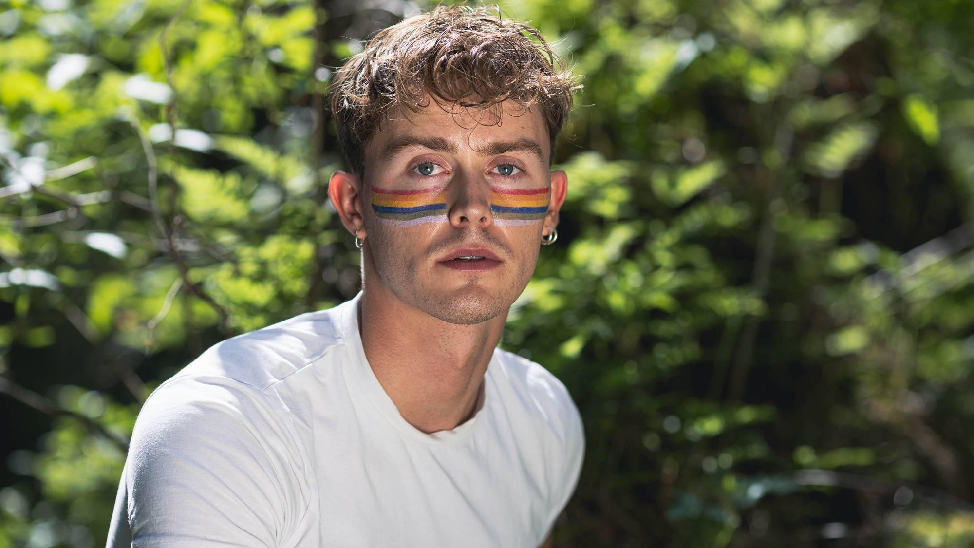 Foto av Jonas. Han har lysebrunt krøllete hår. I ansiktet er det malt pridestripene på kinnene hans. Han har på seg en hvit tskjorte.