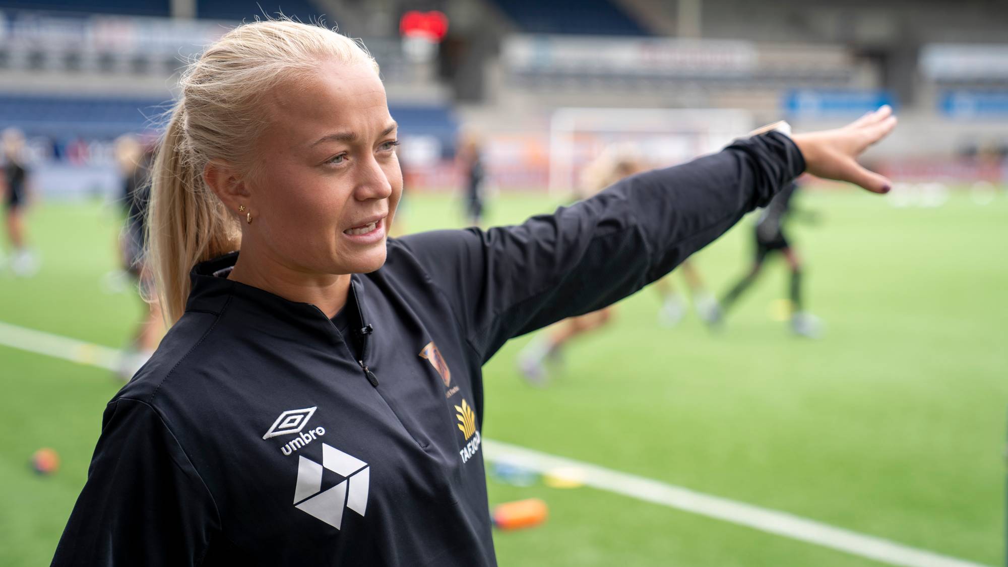 Mette Osvold Muri står på Color Line Stadion og peikar ut på grasmatta, der lagveninnene driv trening.