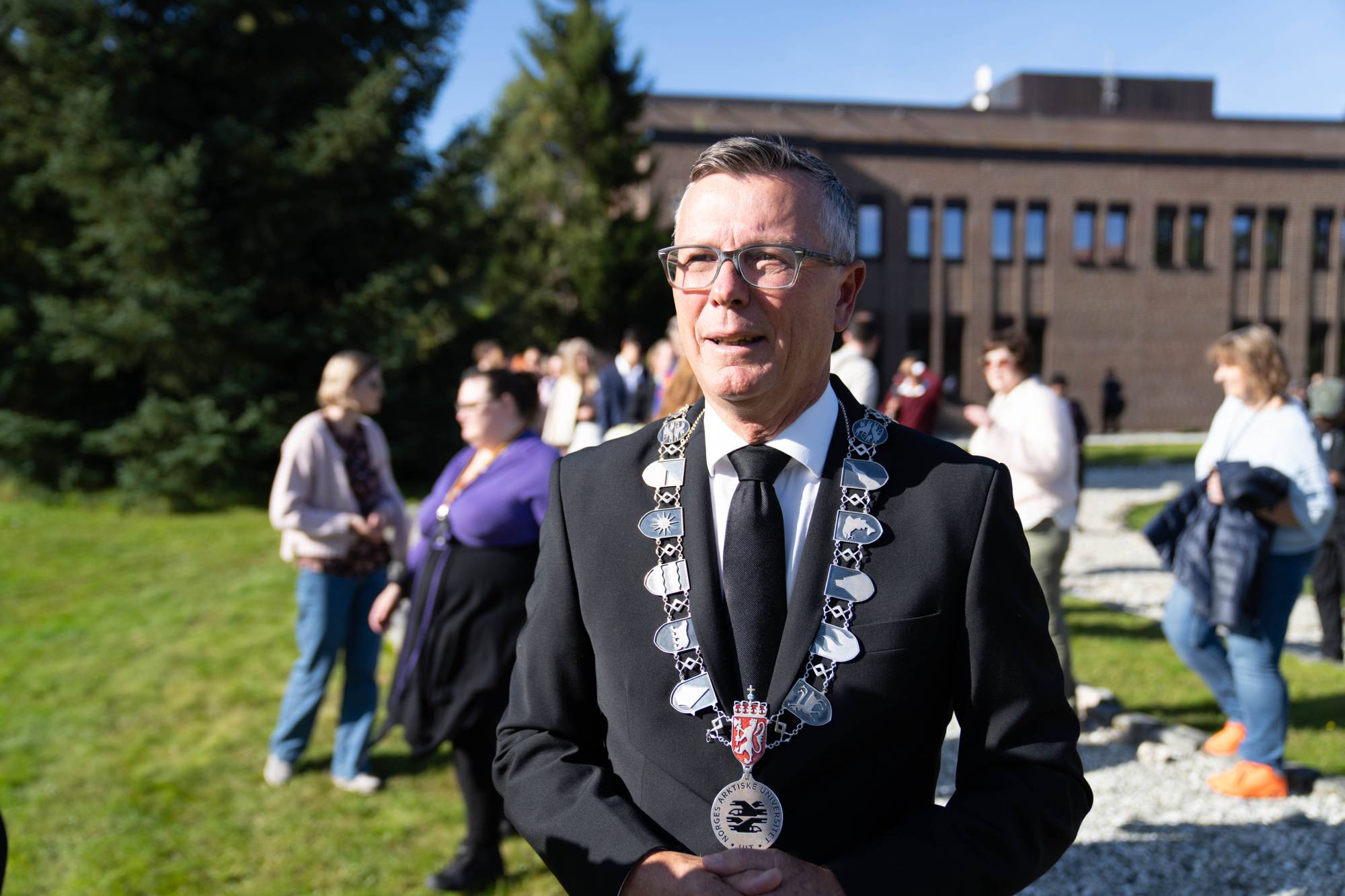 Rektor Dag Rune Olsen med universitetets kjede utendørs foran en murbygning med folk i bakgrunnen. (Bildebeskrivelsen er laget av en KI-tjeneste)
