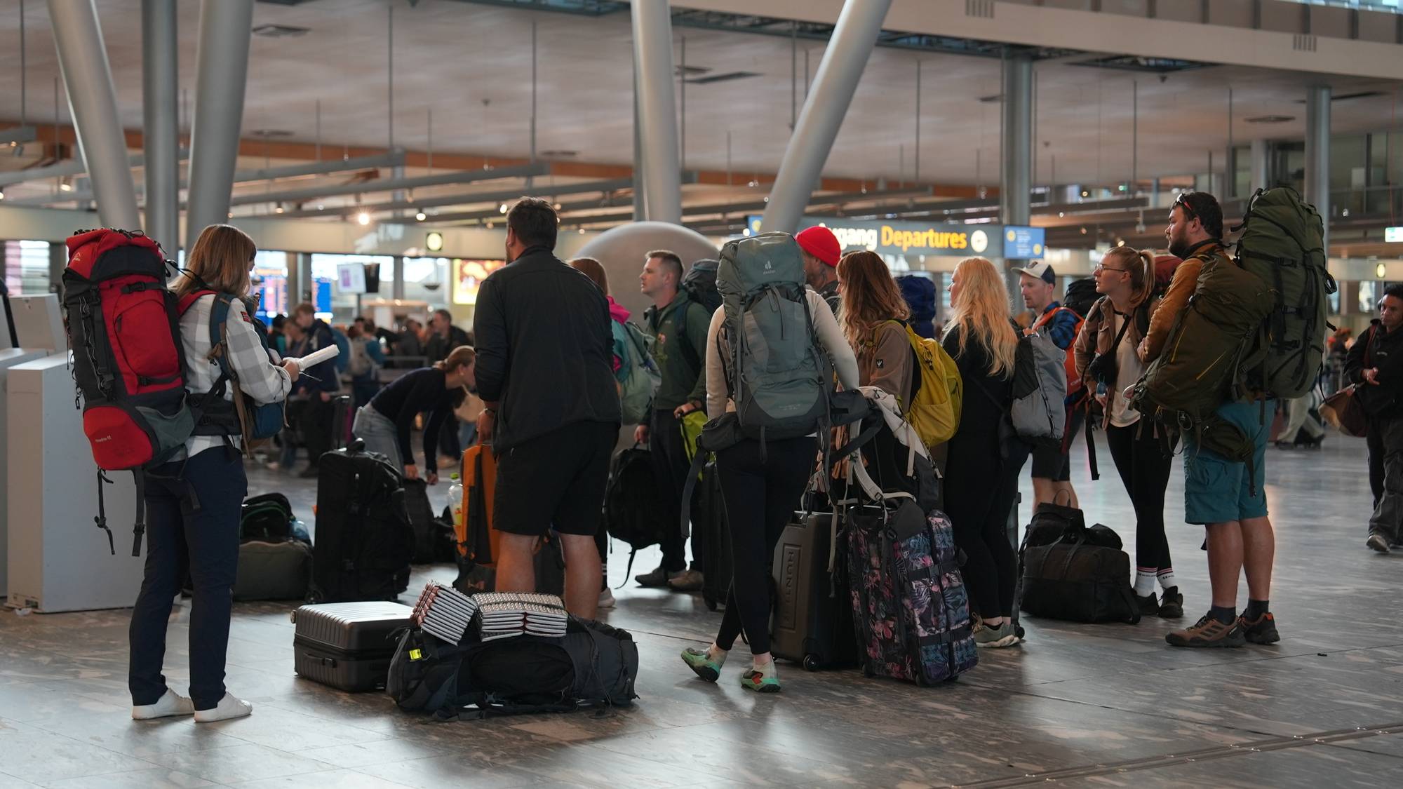 Reisende med store ryggsekker og bagasje står i kø i en flyplasshall. (Bildebeskrivelsen er laget av en KI-tjeneste)