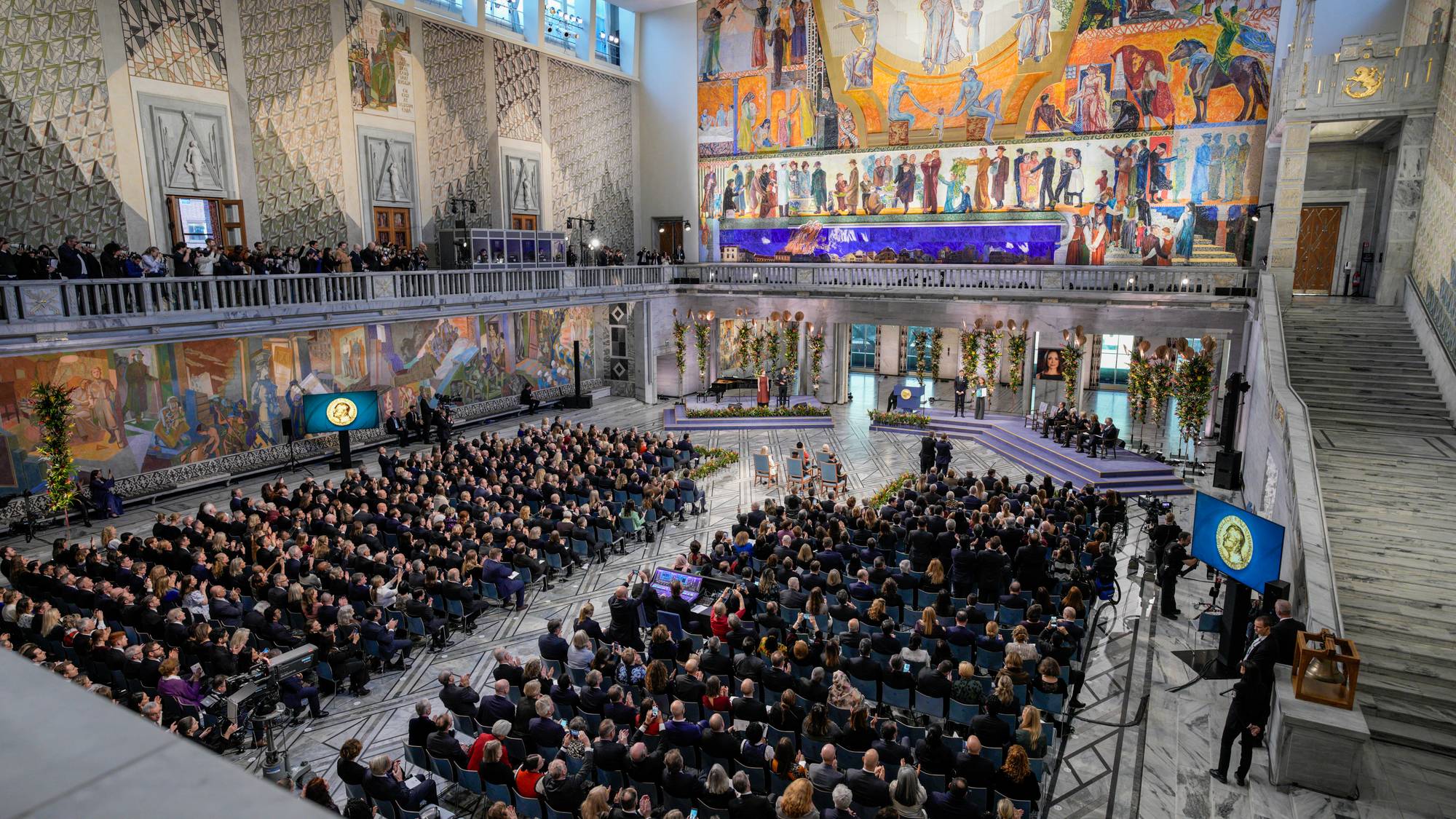 Publikum i Oslos rådhus under nobelseremonien. Storsalen er fylt med folk, dekorert med store malerier. (Bildebeskrivelsen er laget av en KI-tjeneste)