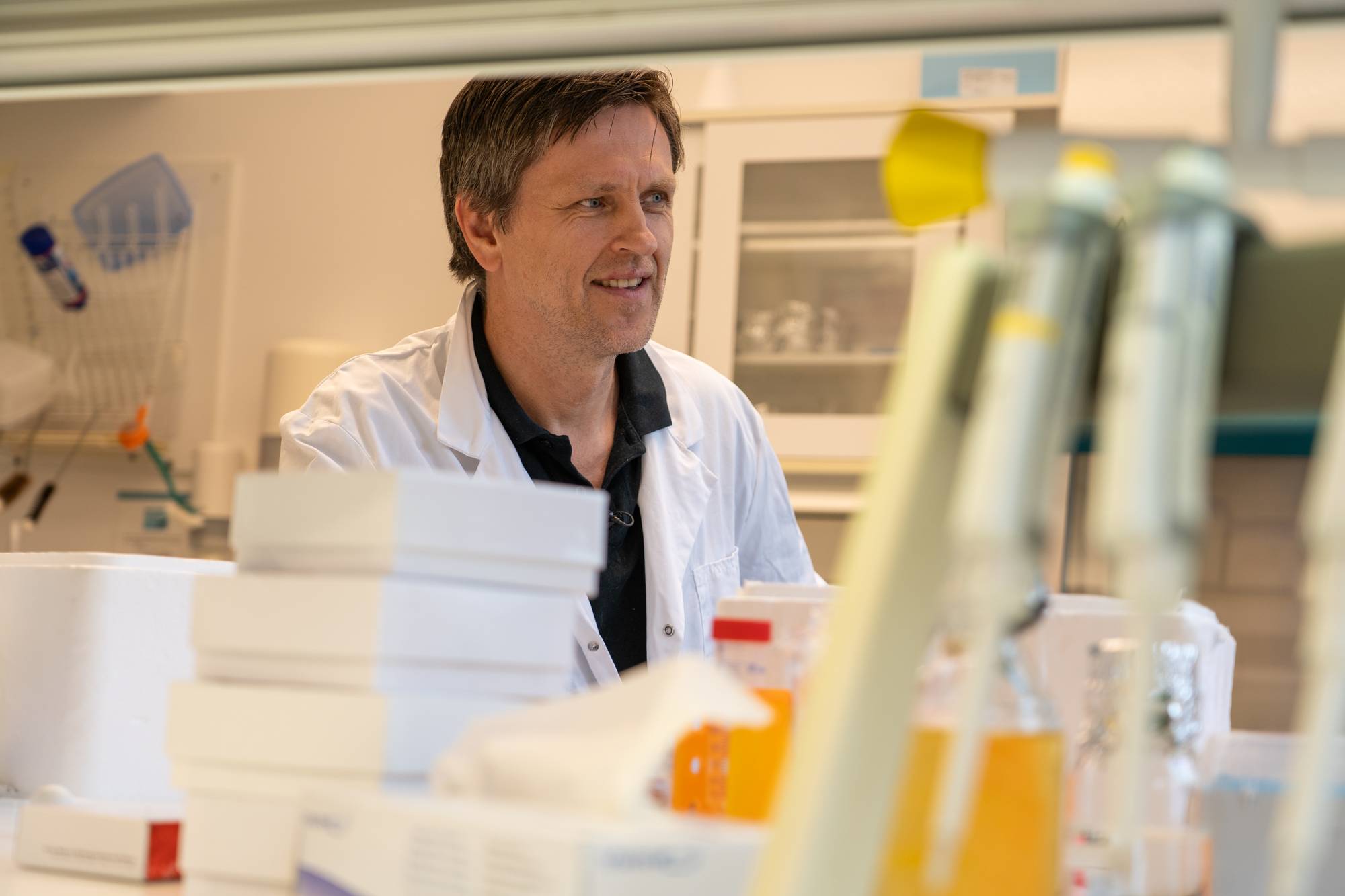 Mann i hvit labfrakk står på et laboratorium med utstyr i forgrunnen. (Bildebeskrivelsen er laget av en KI-tjeneste)