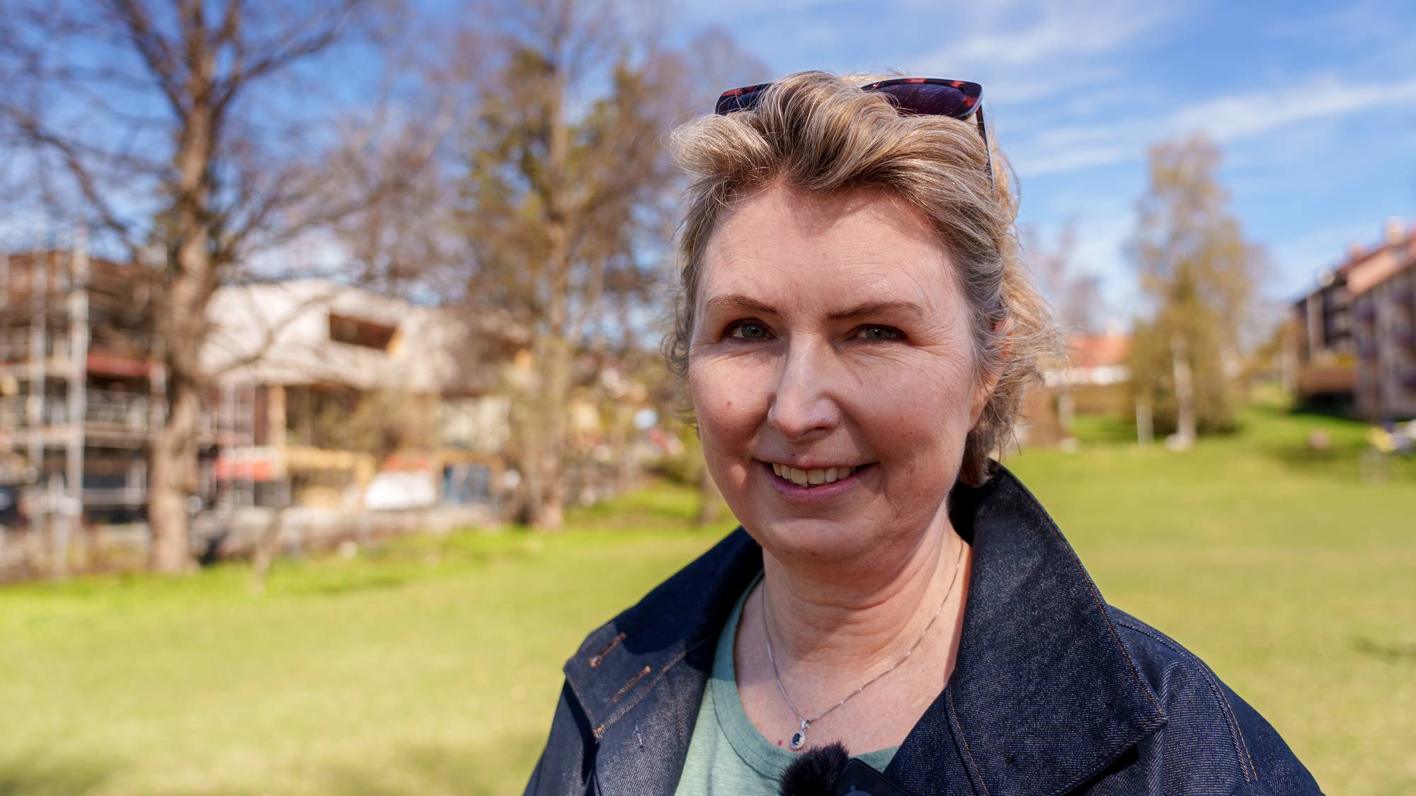 En kvinne smiler mot kameraet. Hun har på seg en grønn genser og en dongerijakke. Bak henne er et grønt område med trær og bygninger. Solen skinner på himmelen. (Bildebeskrivelsen er laget av en KI-tjeneste)