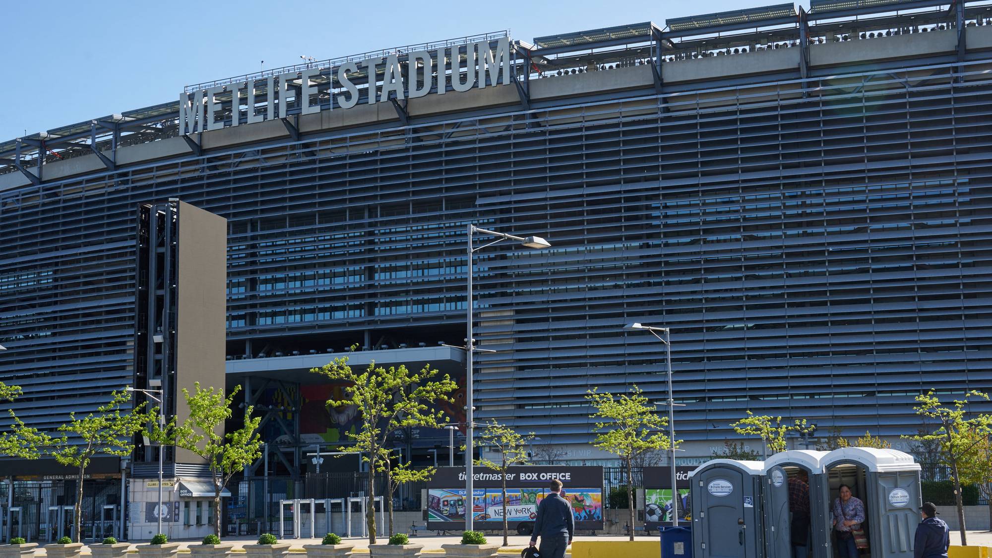 MetLife Stadium står i bakgrunnen. En mann med bag går forbi en rekke utedoer. Det er en bil med billetter. (Bildebeskrivelsen er laget av en KI-tjeneste)
