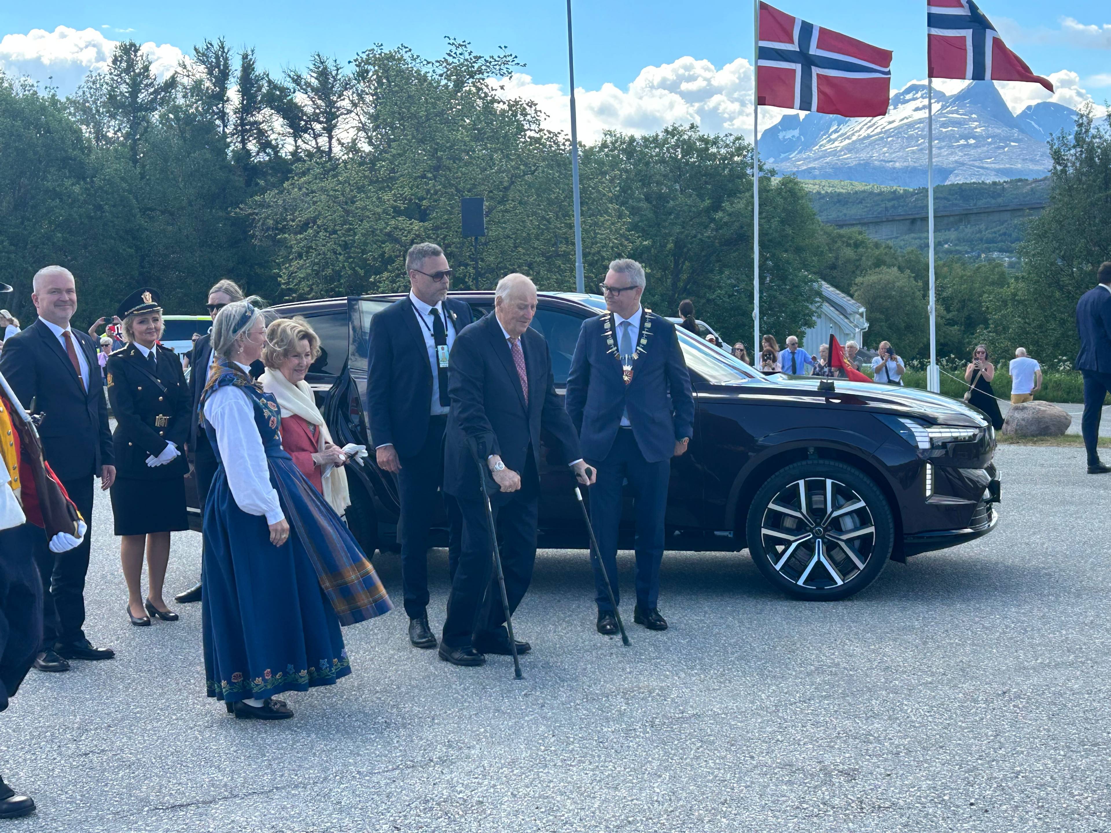 Kong Harald ankommer Saltstraumen på krykker. Dronning Sonja er også på bildet. Bak dem vaier norske flagg i vinden.