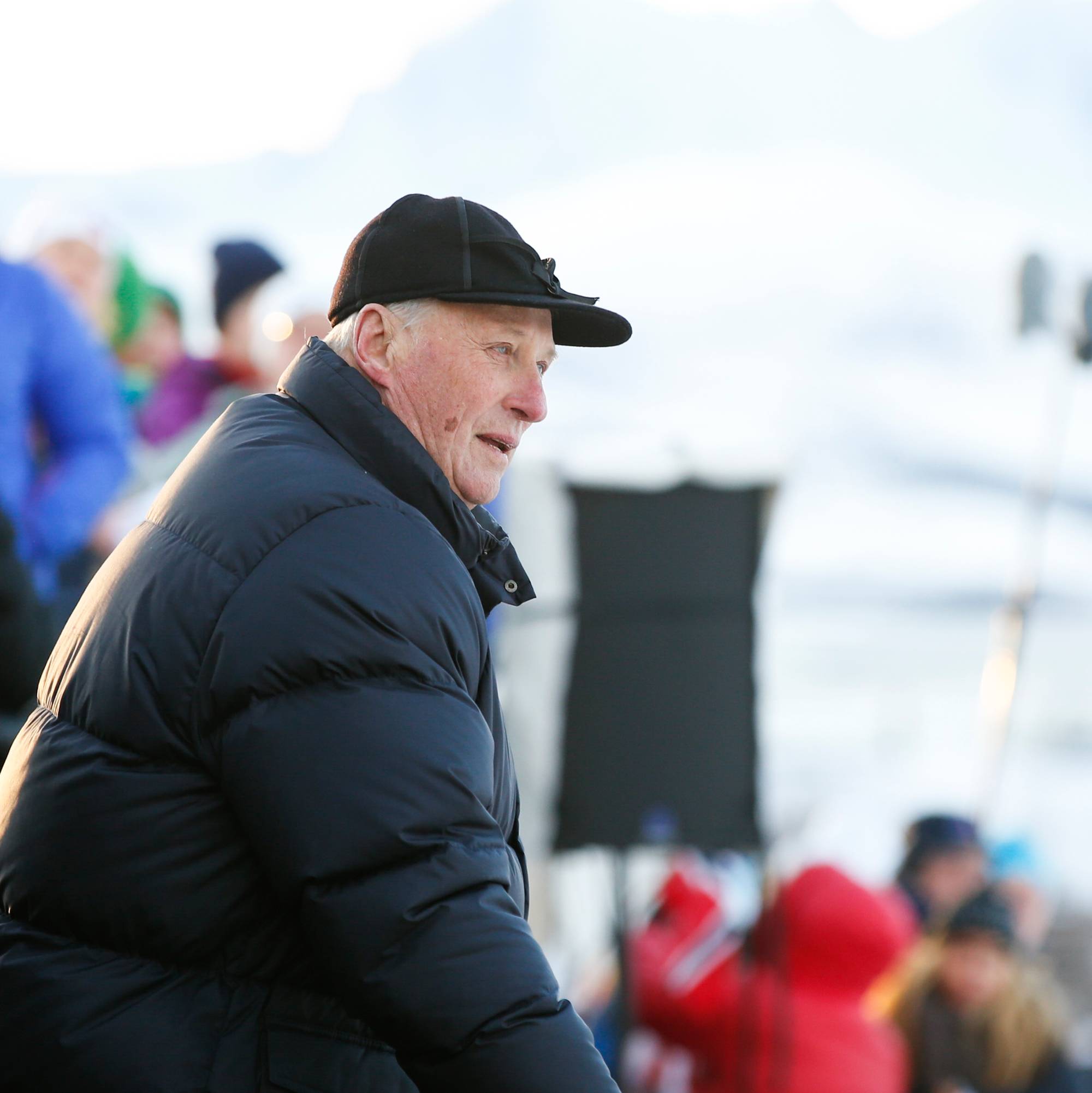 Kong Harald ser mot en skiarena utendørs. Publikum er synlig i bakgrunnen med snødekt landskap. (Bildebeskrivelsen er laget av en KI-tjeneste)
