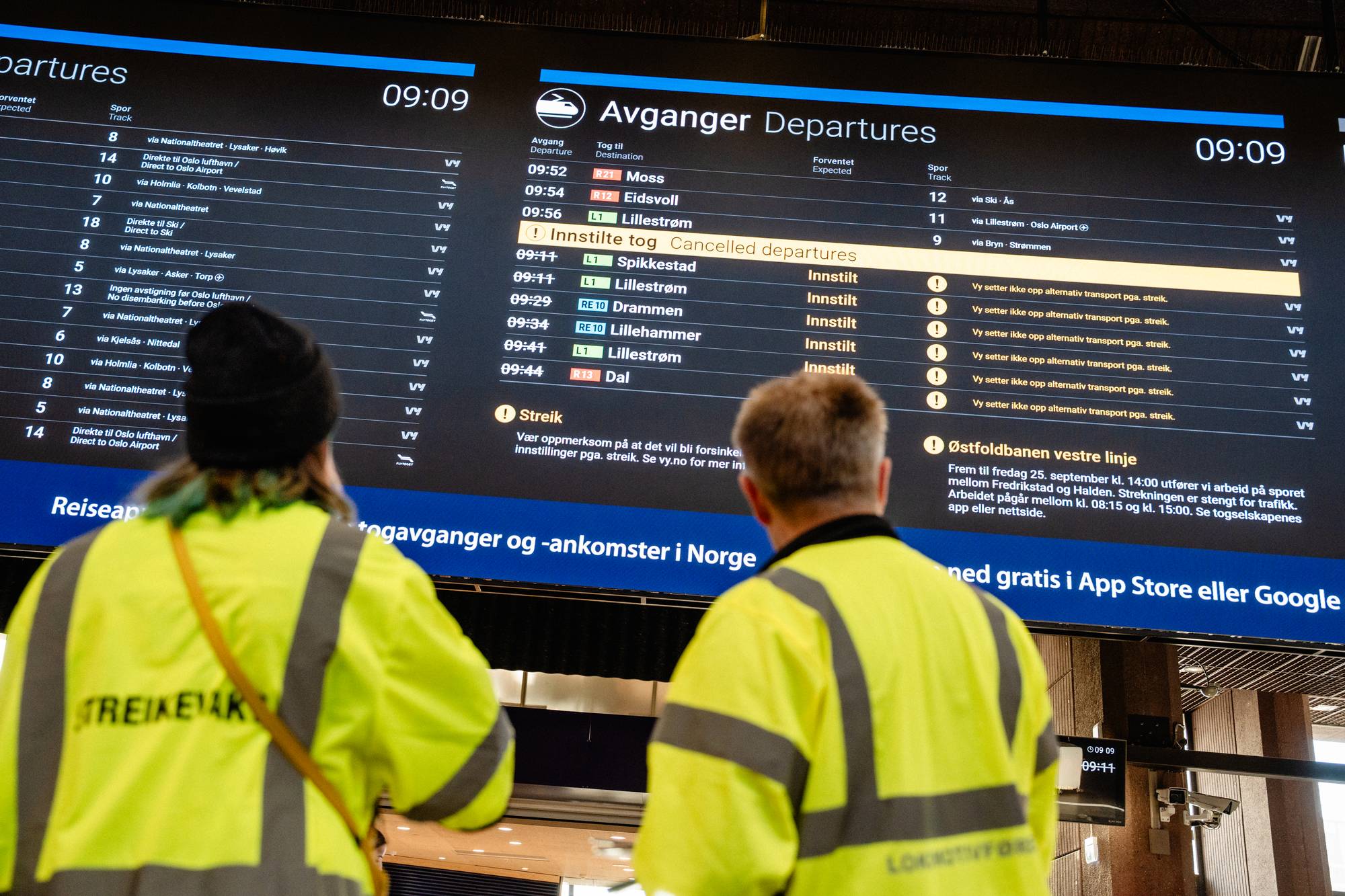 To personer i gul refleksvest ser på en digital informasjonstavle med innstilte togavganger. (Bildebeskrivelsen er laget av en KI-tjeneste)