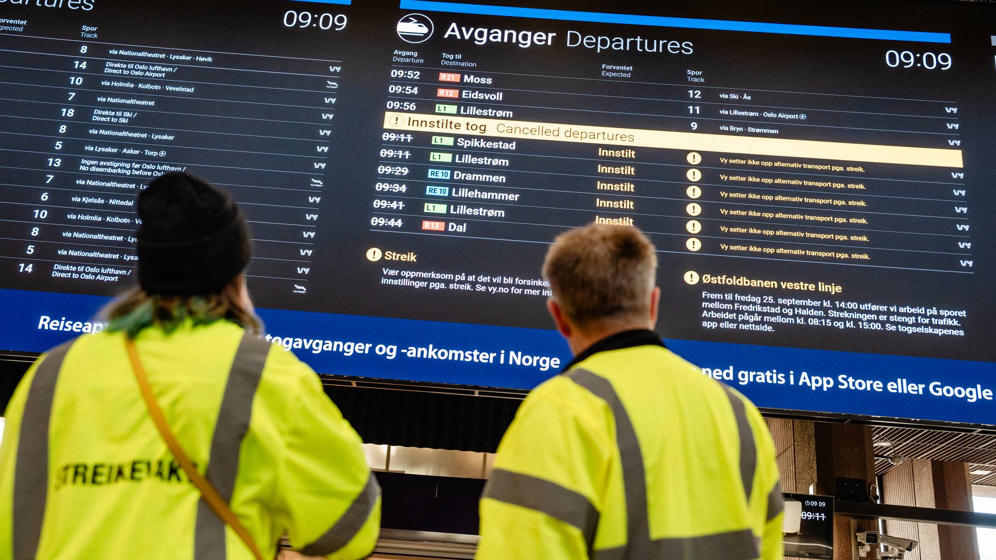 To personer i gul refleksvest ser på en digital informasjonstavle med innstilte togavganger. (Bildebeskrivelsen er laget av en KI-tjeneste)